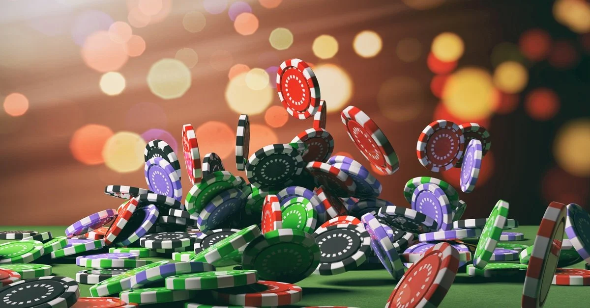 Red, black, purple, and green poker chips fall onto a green felt table. Blurred lights twinkle in the background.