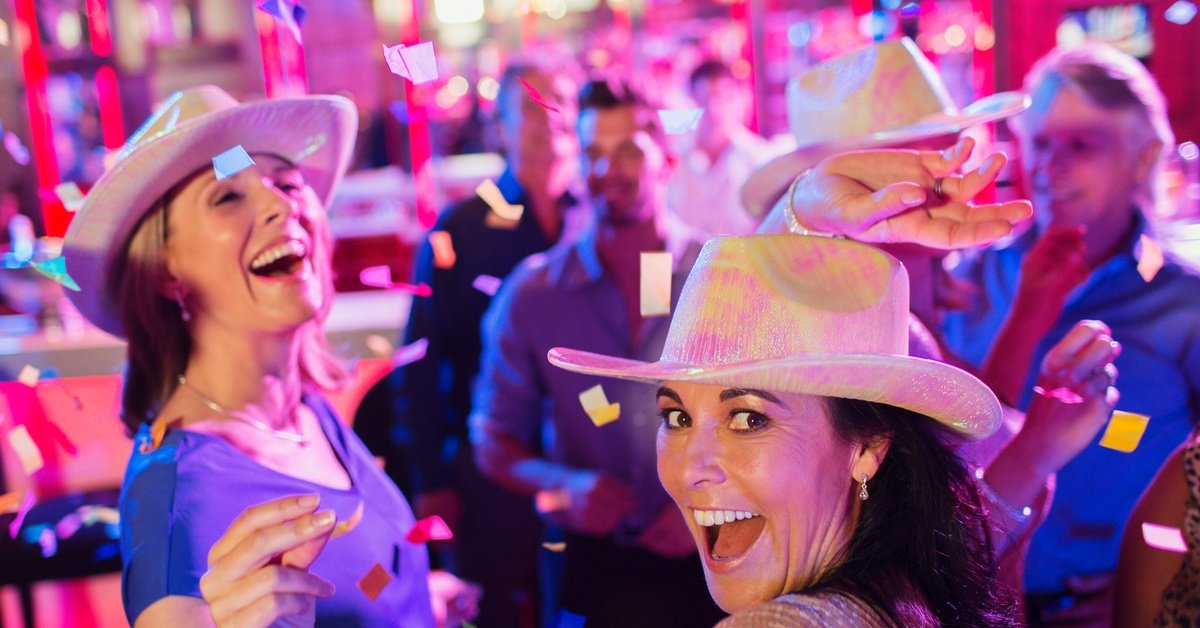 People celebrate under neon pink lights and a shower of confetti. Some wear iridescent white-and-pink cowboy hats.