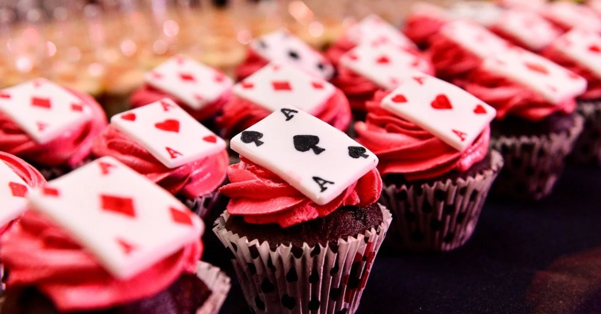 Rows of chocolate cupcakes with red frosting are on a table. Each cupcake is topped with a fondant playing card.