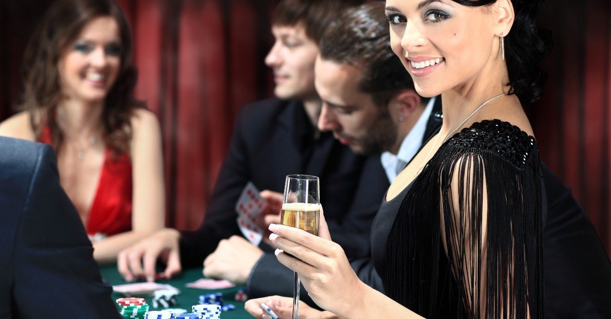A group of dressed-up adults is at a table with playing cards and poker chips. One woman holds a champagne flute.