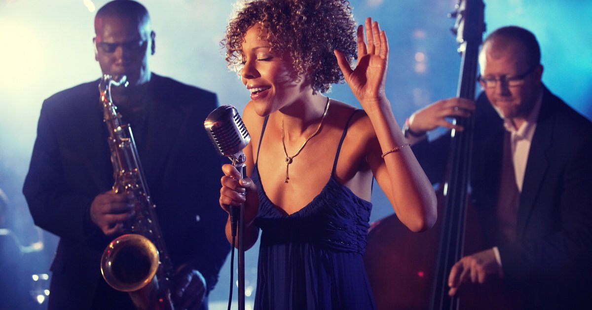A singer sings into a vintage microphone while raising one hand. A saxophone player and a bassist perform behind her.