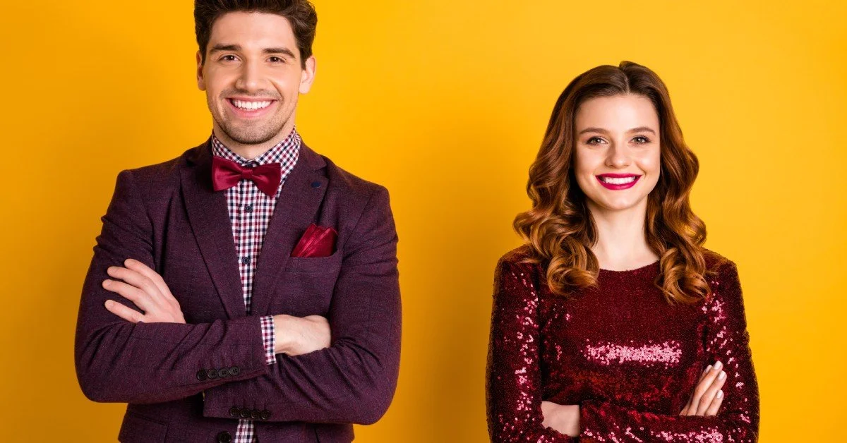 A smiling man and woman stand side by side. He wears a brown jacket and red bow tie, and she wears a red sequin dress.