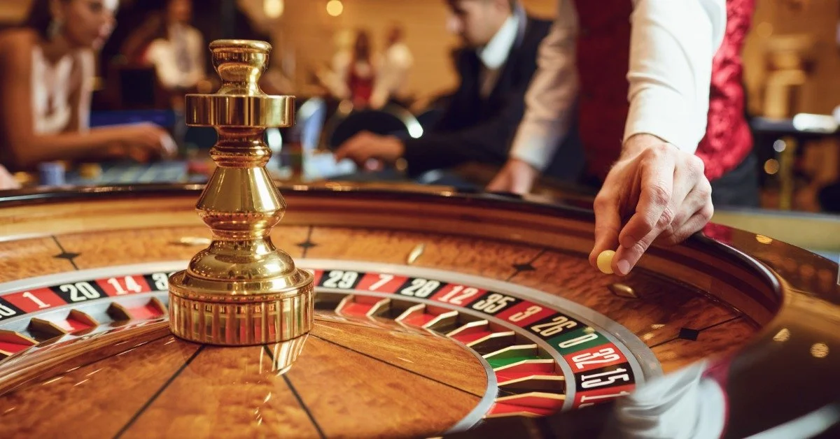 A dealer places a small white ball in a roulette wheel with numbered slots. Blurred players sit behind the table.