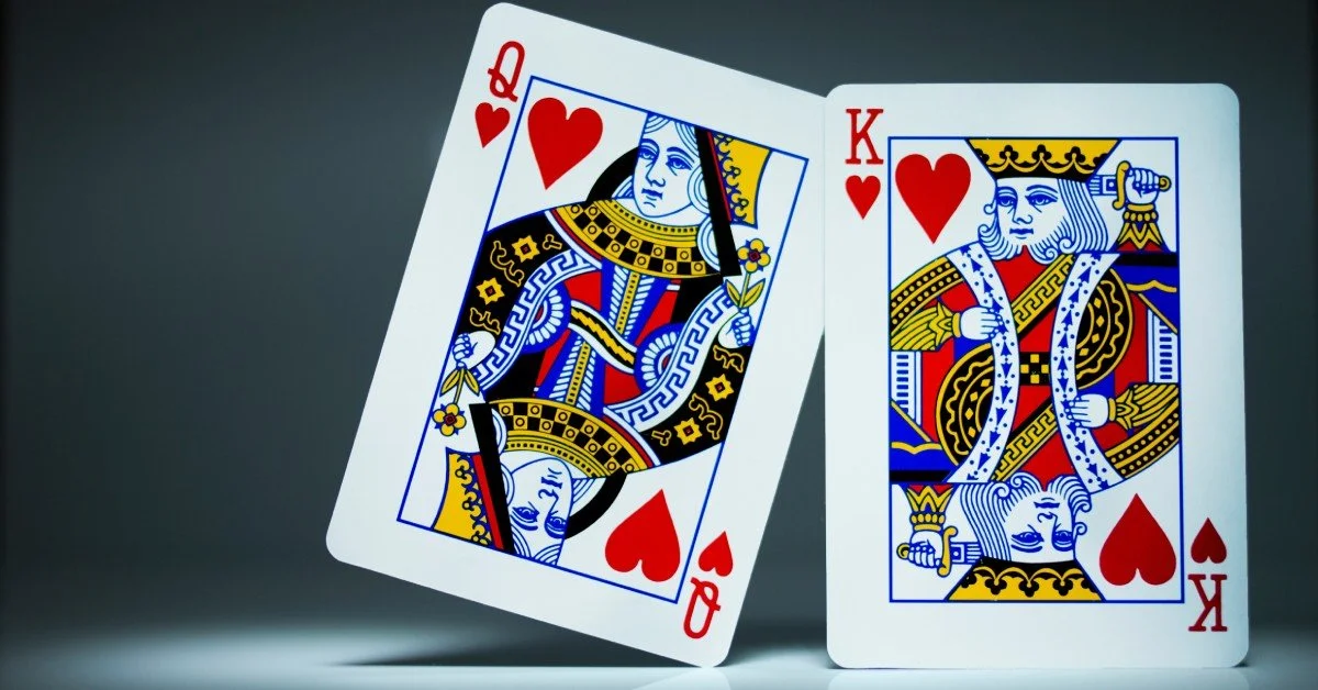 A queen of hearts card is tilted to lean against a king of hearts card. The cards are well-lit before a dark background.