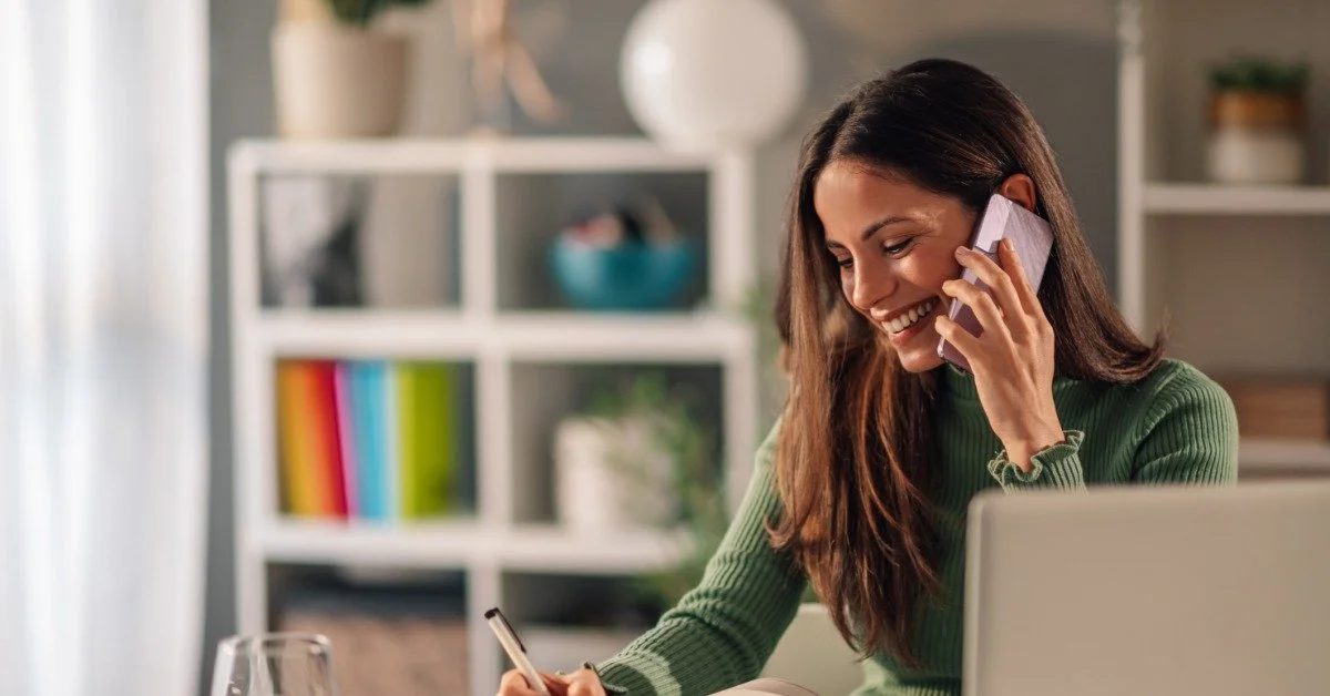 A woman sits at a desk in front of a laptop. She holds a phone to her ear and smiles as she writes in a notebook.