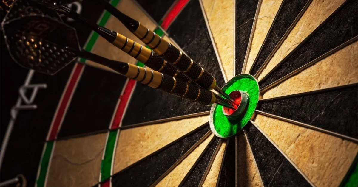 A close-up of a dartboard shows alternating black and beige sections. Three darts are embedded in the red bullseye.