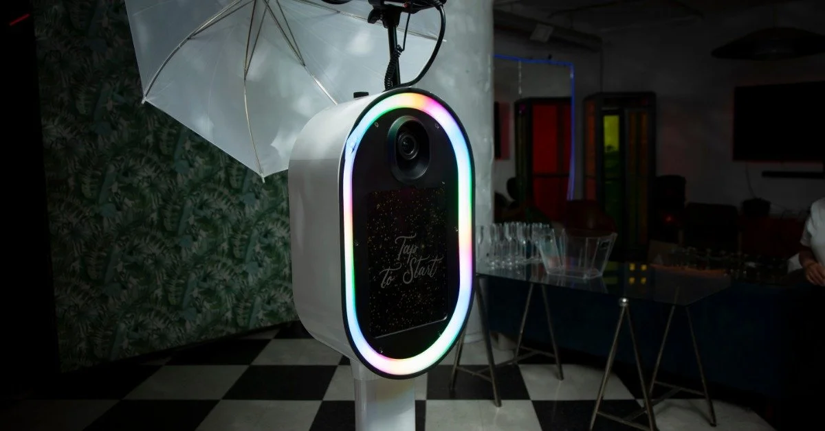 A white photo booth kiosk with a glowing multicolor light ring stands in front of a large photography umbrella.