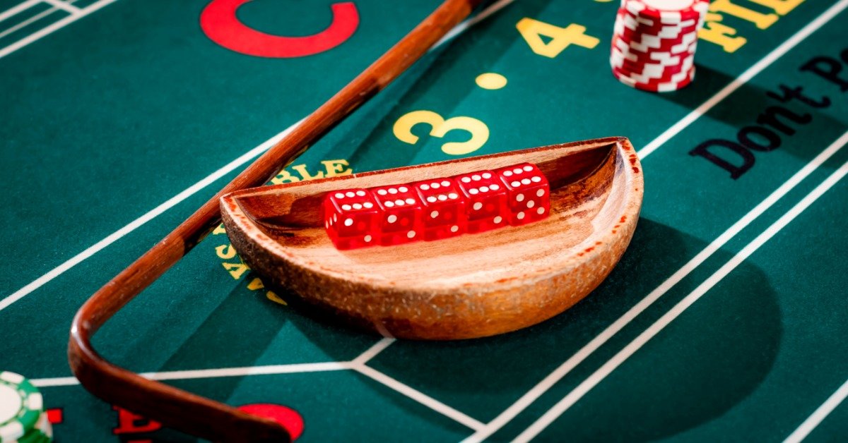 Red dice in a wooden bowl, stacks of red and green poker chips, and a craps dice stick are on a green craps table.