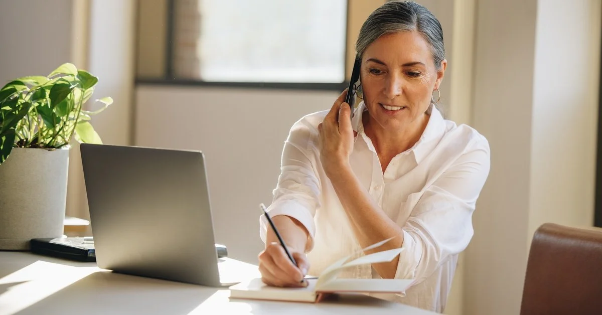 A smiling woman sits at a desk with a laptop. She smiles and writes in a notebook while listening to her cell phone.