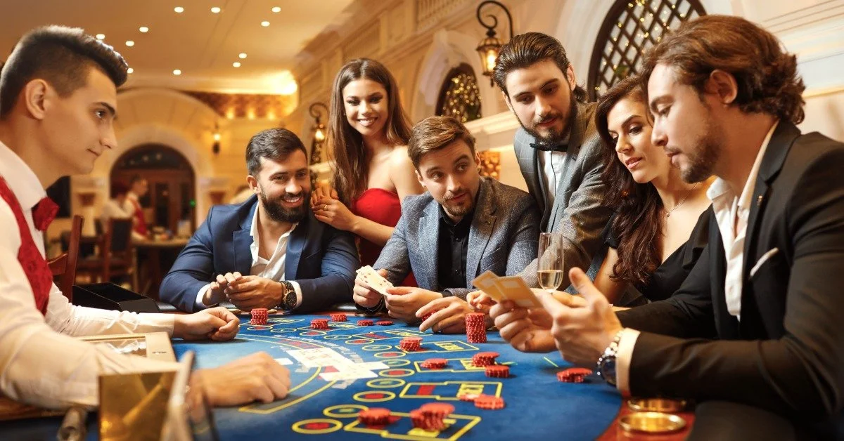 A man in a black suit studies his cards while other guests watch around the casino table. A dealer sits opposite them.