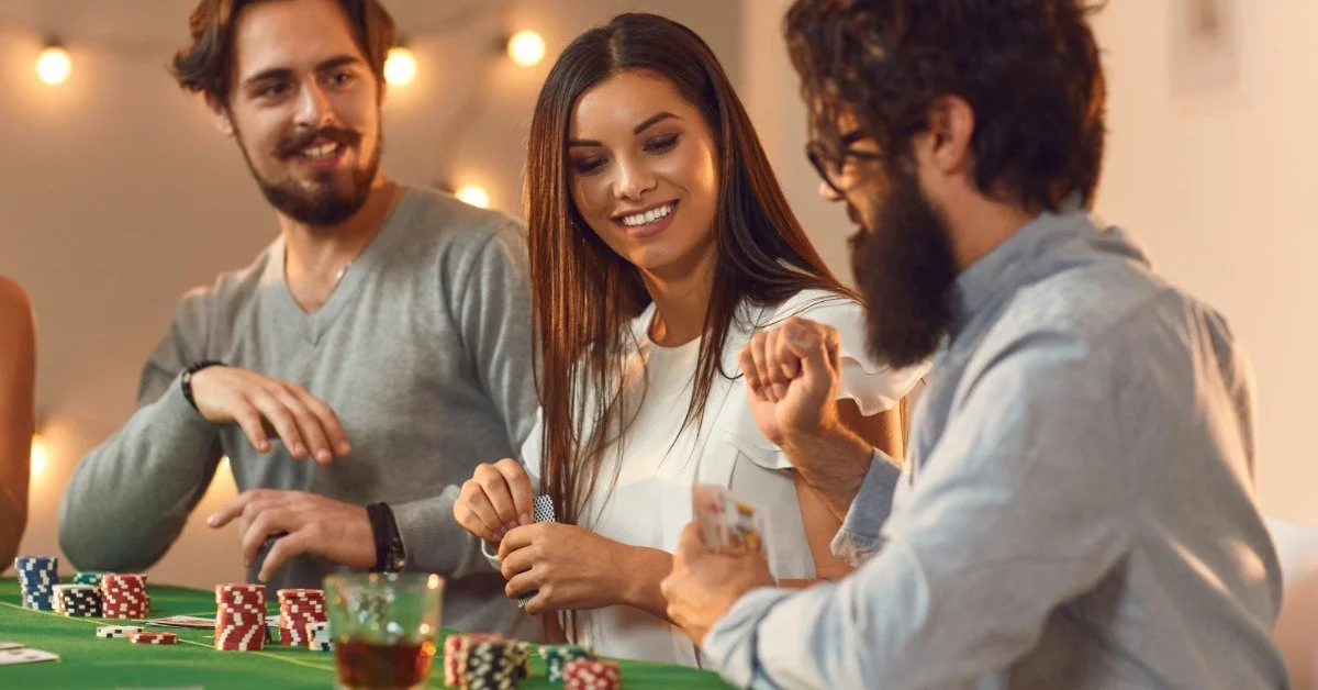 A small group plays cards at a table covered with green felt. Red, white, black, and green poker chips are on the table.