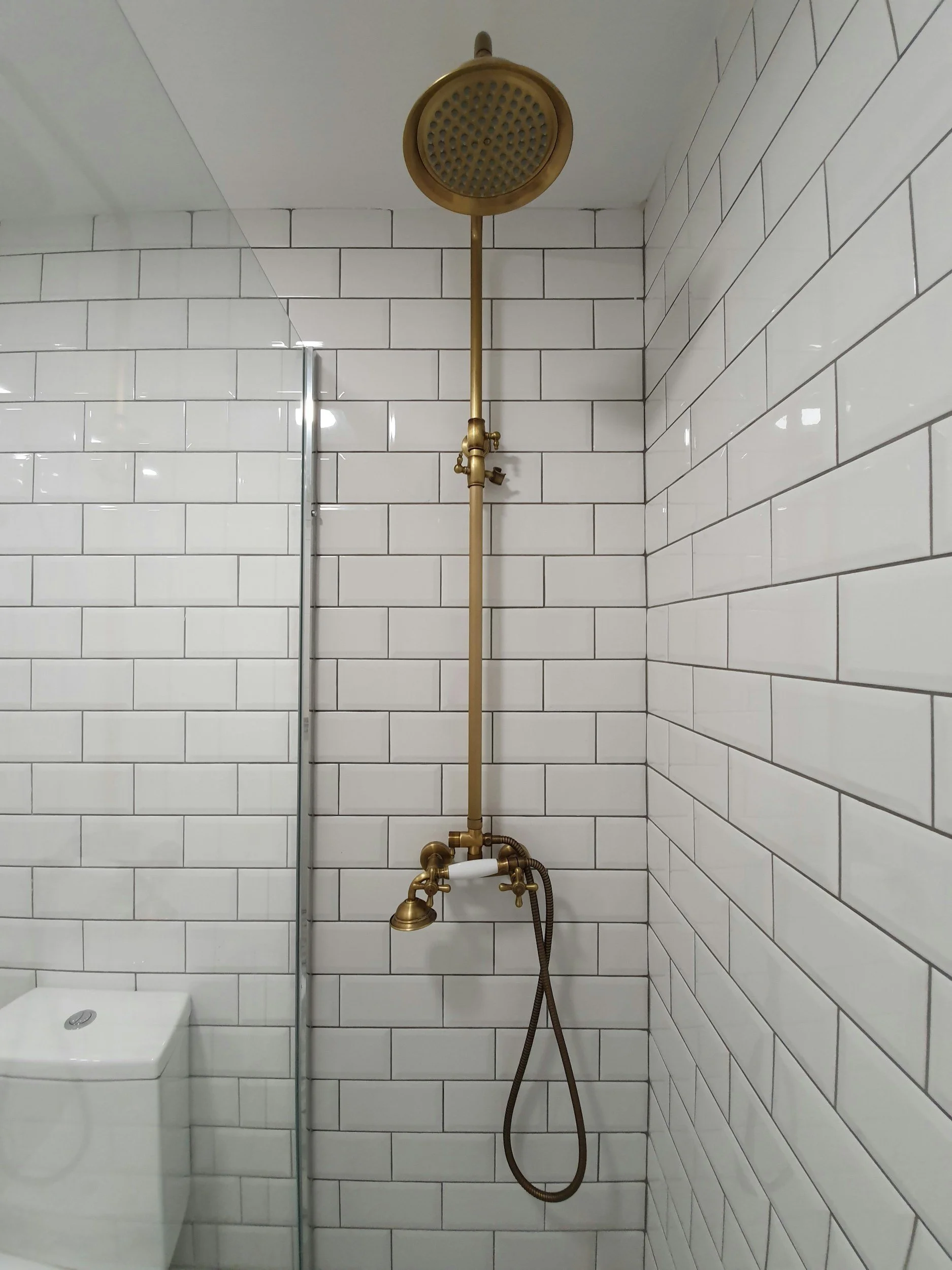 Bathroom shower with a gold-colored overhead showerhead, brass fixtures, a handheld shower wand, white subway tile walls, and a partial view of a toilet.
