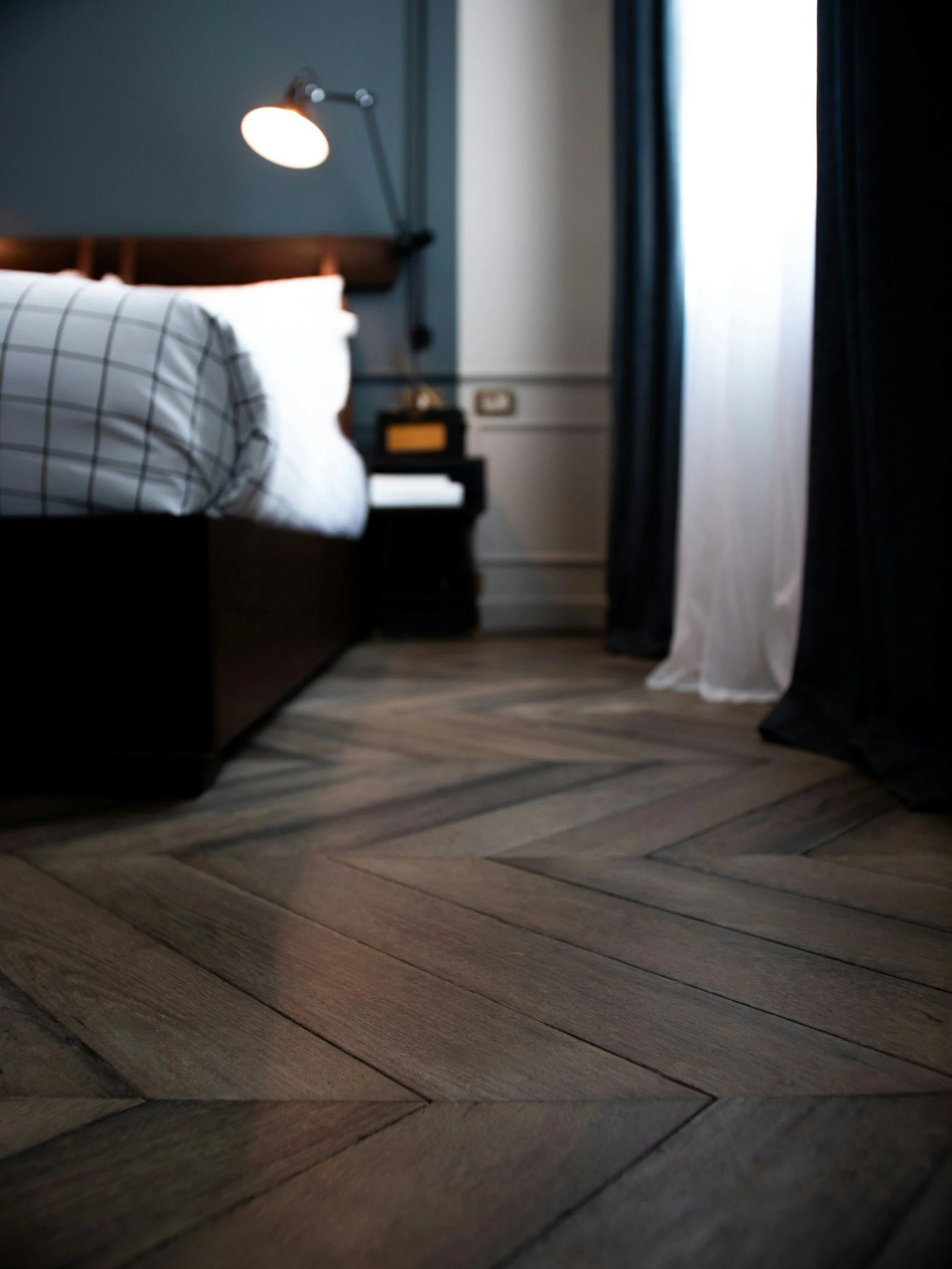 A bedroom with a bed, checkered white and black pillows, a bedside table with books, a wall-mounted lamp, and curtains, with a wooden floor arranged in a herringbone pattern.