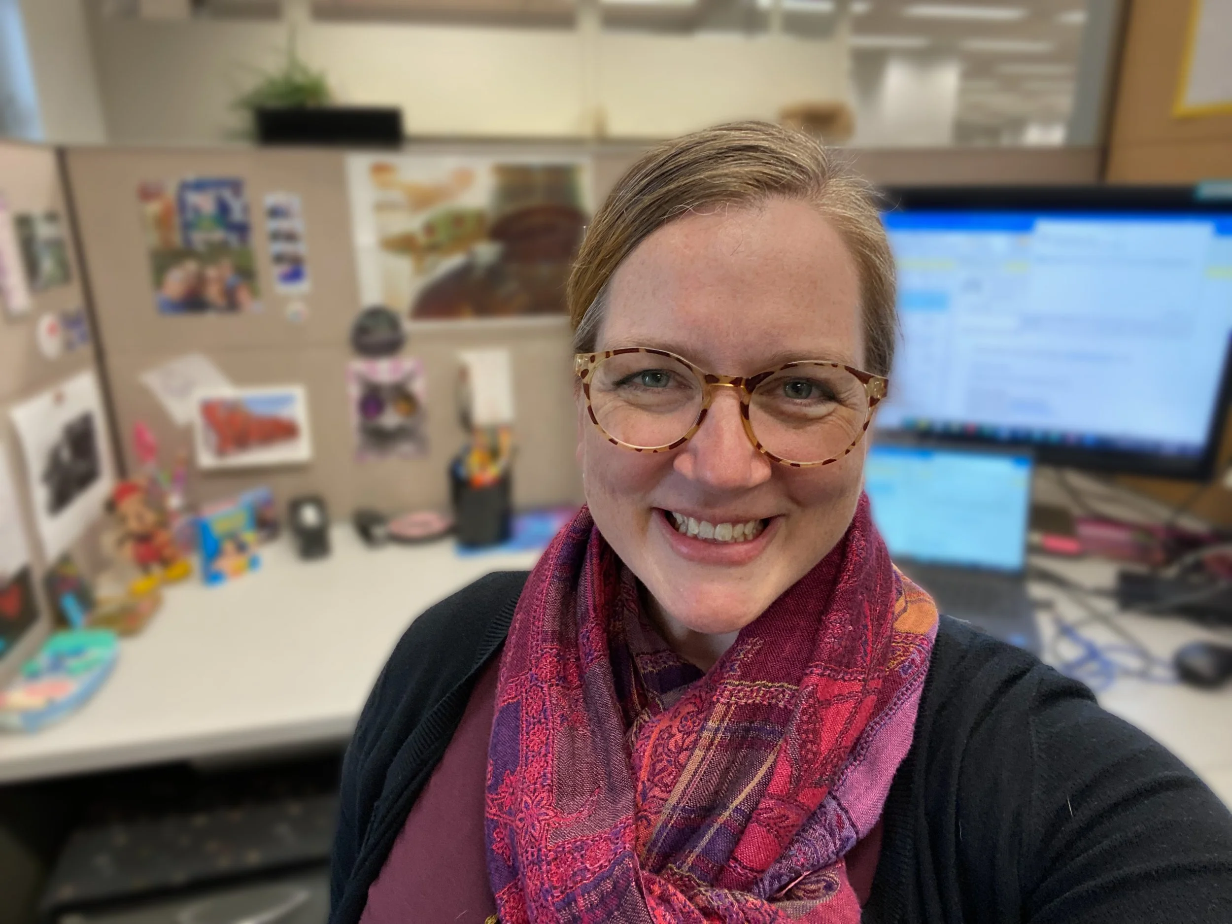Smiling woman with glasses and a red and purple scarf in an office with cubicles and computer monitors in the background.