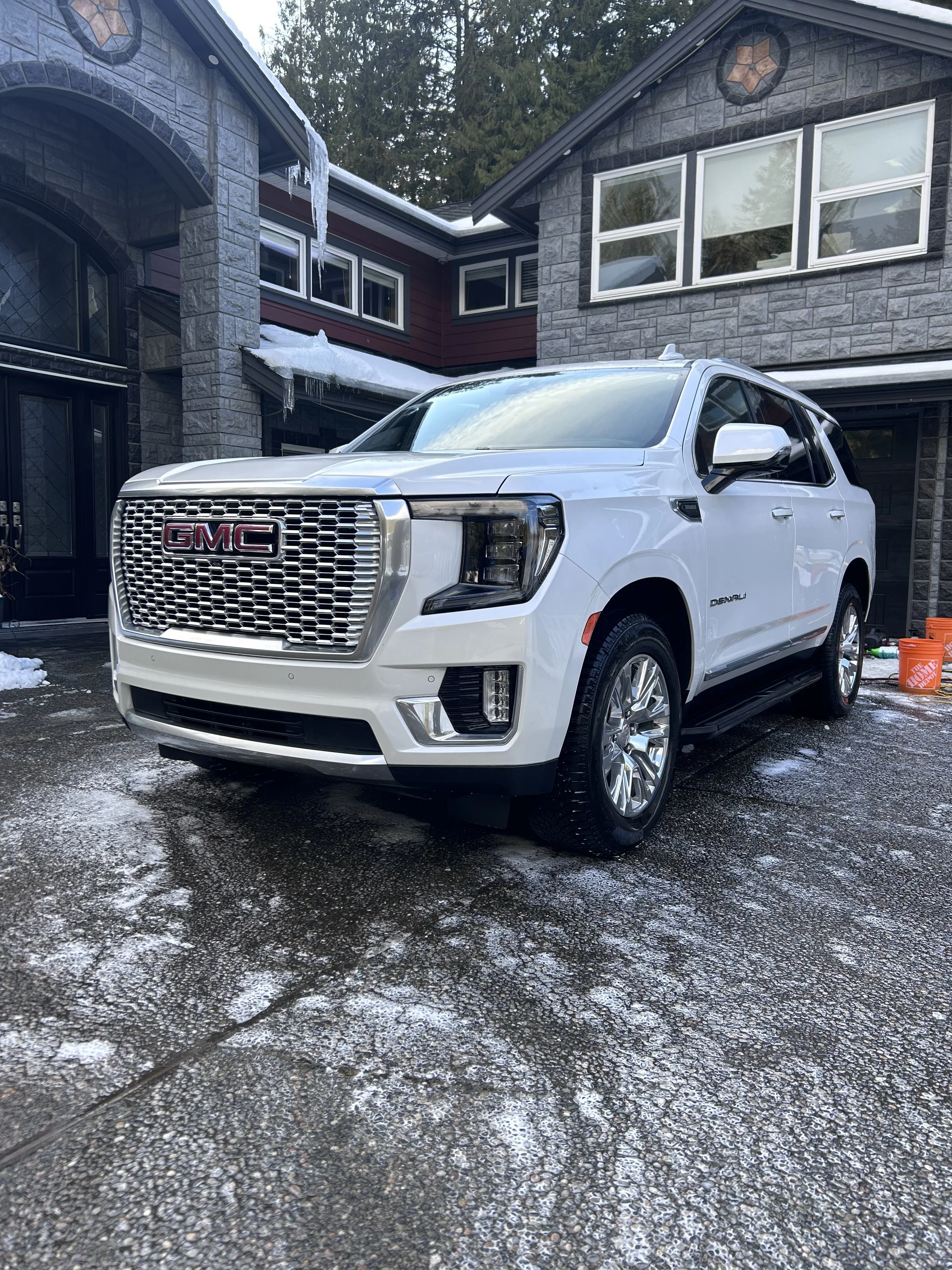White GMC Denali SUV parked in a driveway in front of a stone house with snow and ice around.