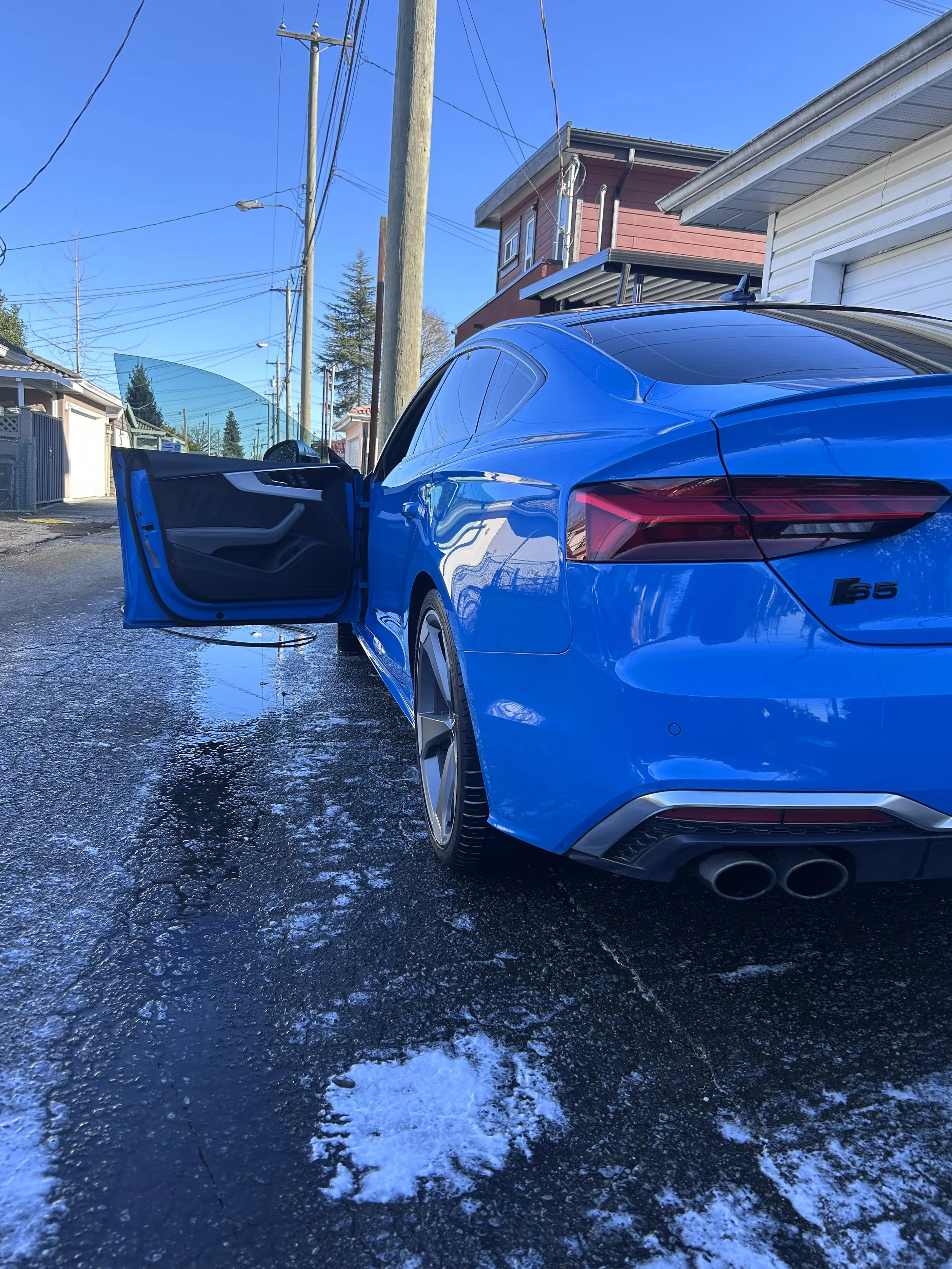 Blue sports car with an open door on a wet, icy road near residential buildings, under a clear sky.