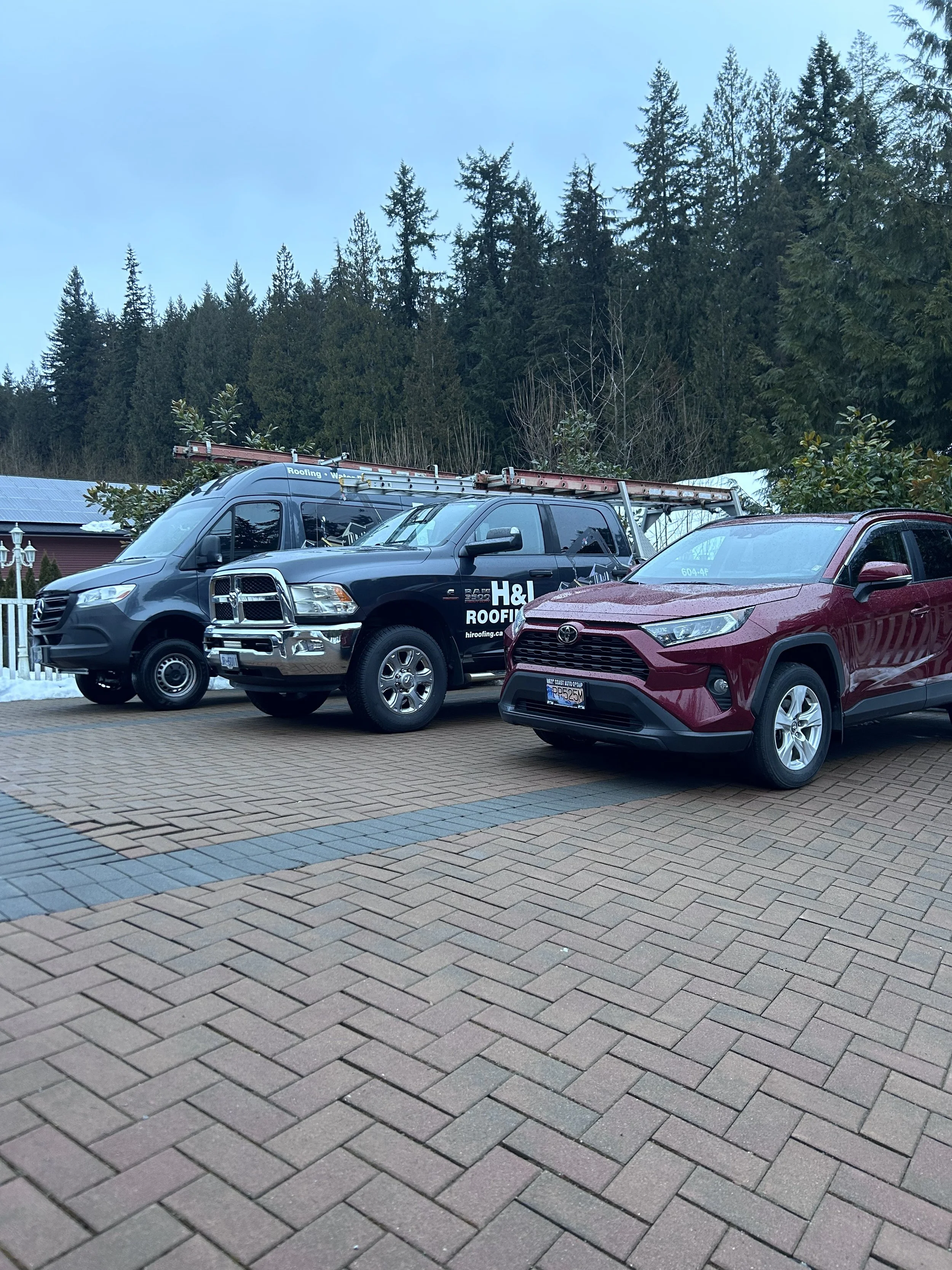 A parked black roofing van with a ladder, a dark gray truck with "H&I Roofing" signage, and a maroon SUV on a paved driveway surrounded by trees.