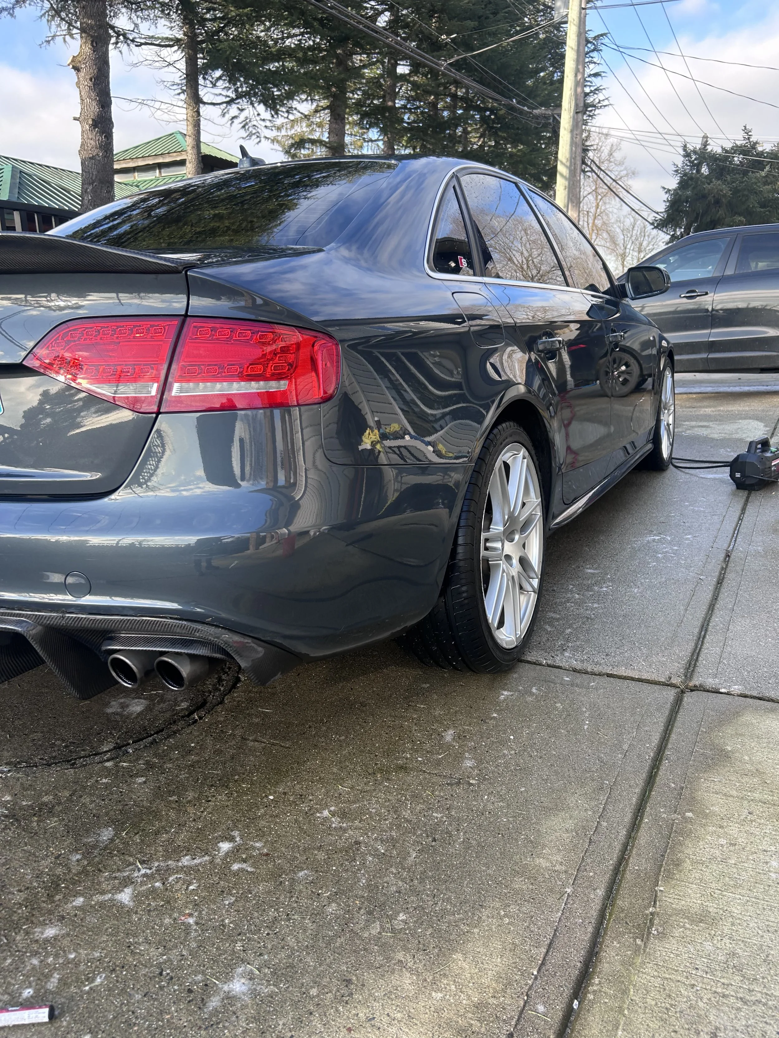 Rear view of a dark-colored sedan with tinted windows, parked on a driveway, trees in the background.