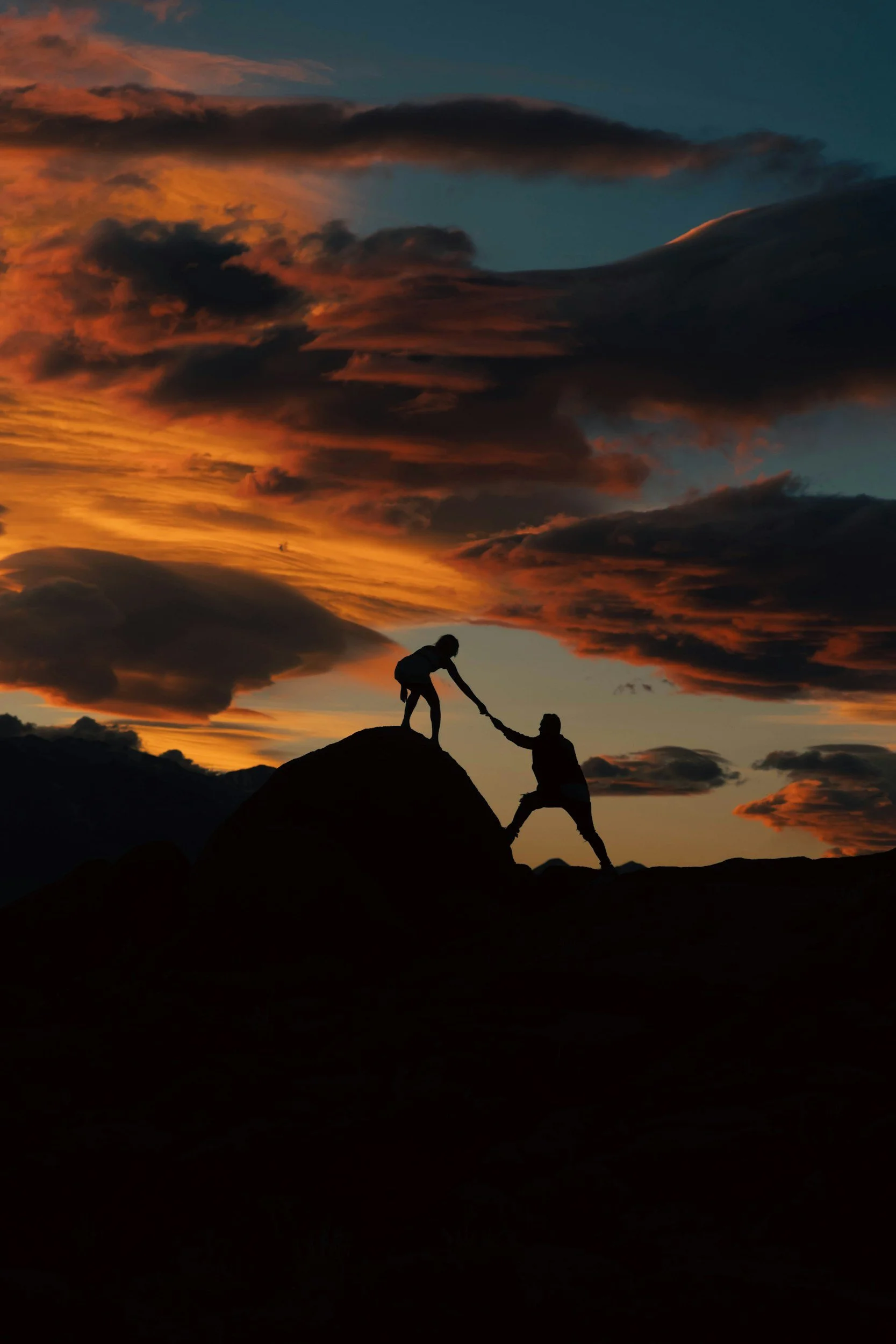 Two people are on a hilltop during sunset with dramatic dark clouds in the sky. One person is helping the other up, creating a silhouette effect against the vibrant sky.