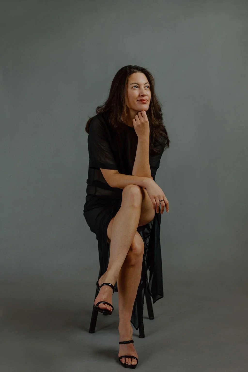 A woman with long dark hair wearing a black dress and high heels, sitting on a stool with one leg crossed over the other, posed thoughtfully against a plain gray background.