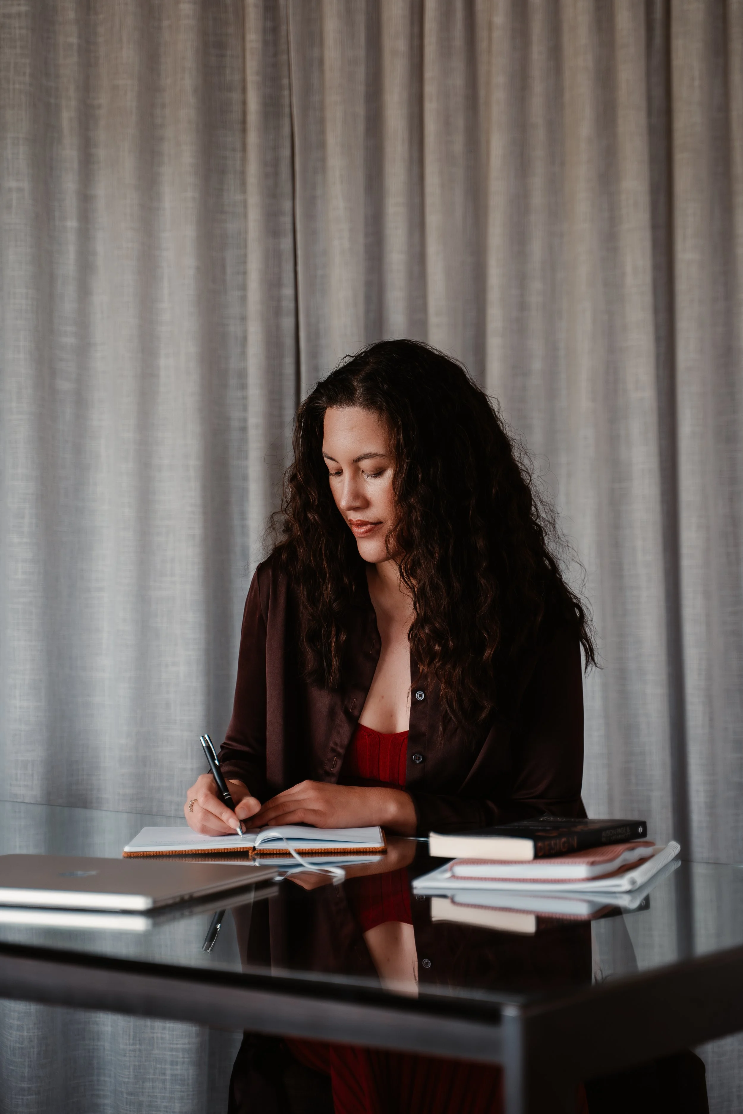 A woman with long curly hair writing in a notebook with a pen, sitting at a reflective black table with books, a laptop, and papers, in a room with gray curtains.