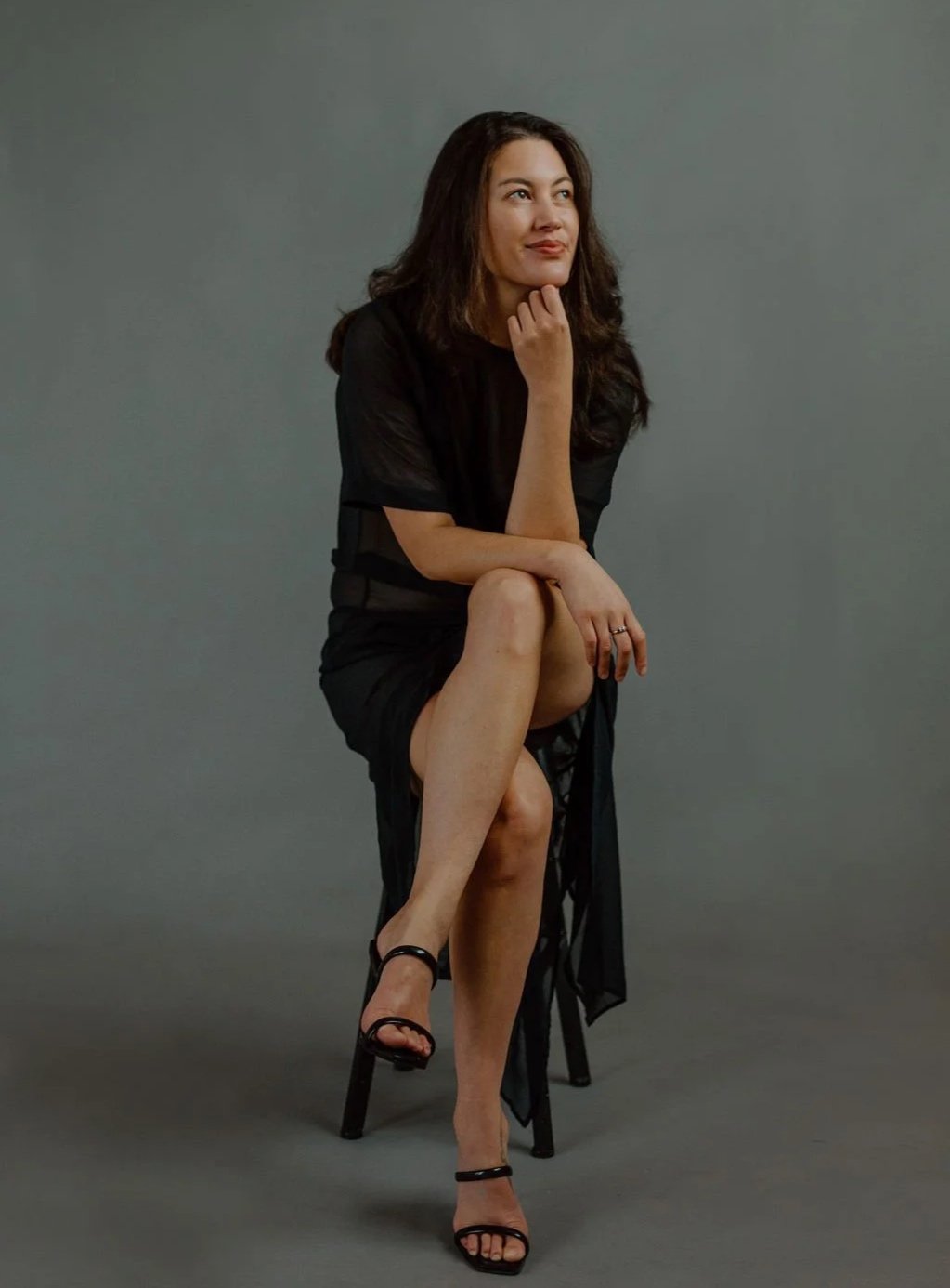 Woman sitting on a black chair, posing with her right ankle resting on her left knee, dressed in a black dress and black high heels, with a thoughtful expression against a plain gray background.