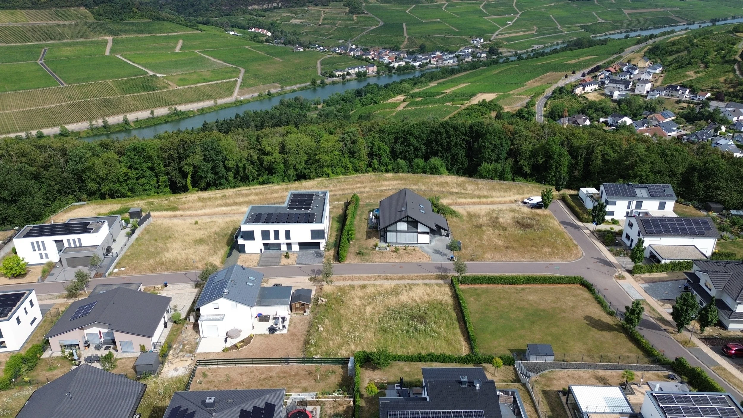 Vue aérienne de maisons modernes avec toits équipés de panneaux solaires, entourées de terrains en pelouse, avec une forêt dense et un fleuve en arrière-plan