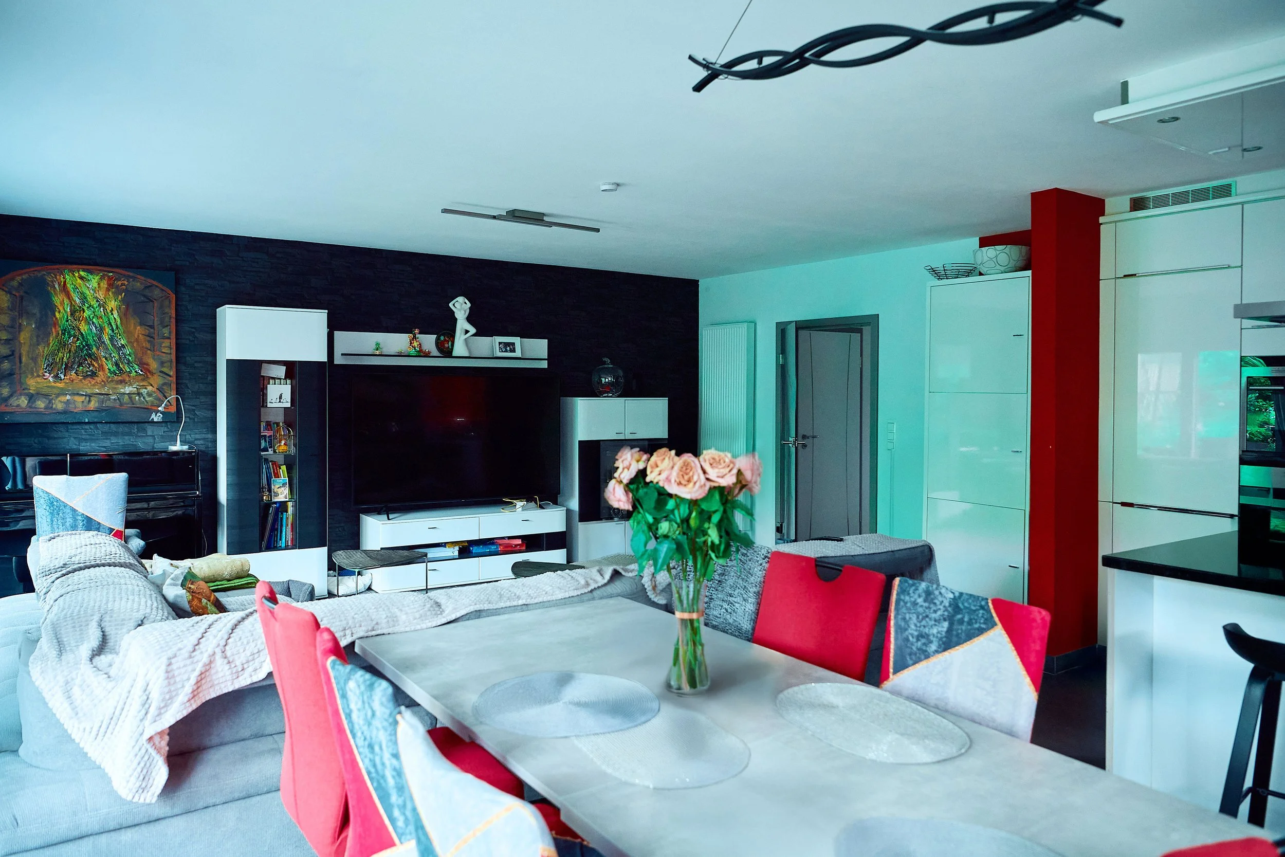 Salon moderne avec un mur noir, un grand téléviseur, une table avec un vase de roses, et une cuisine blanche en arrière-plan.