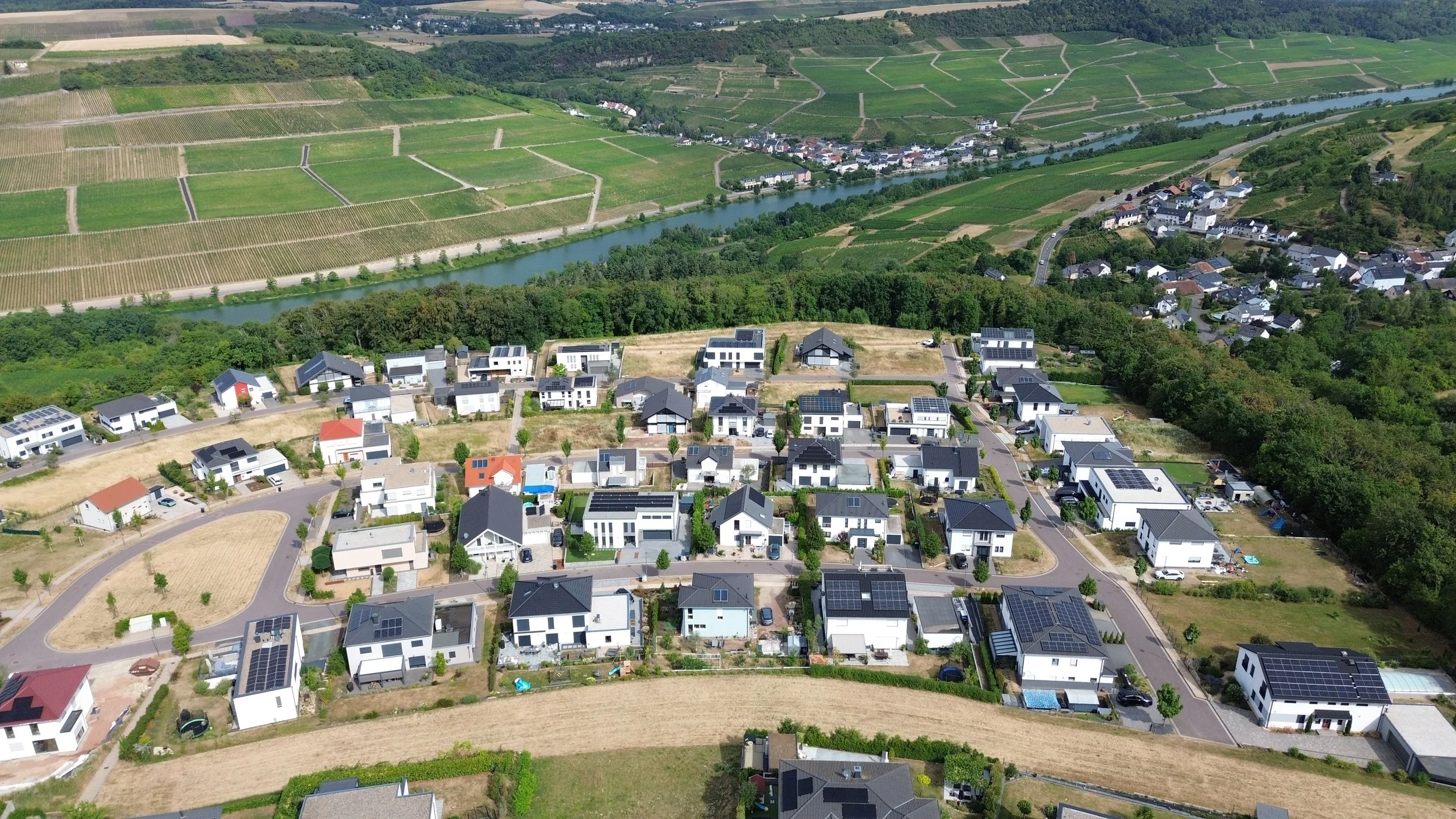 Vue aérienne d'un quartier résidentiel avec des maisons modernes équipées de panneaux solaires, entouré de champs agricoles et de zones verdoyantes, avec une rivière serpentant à travers la scène.