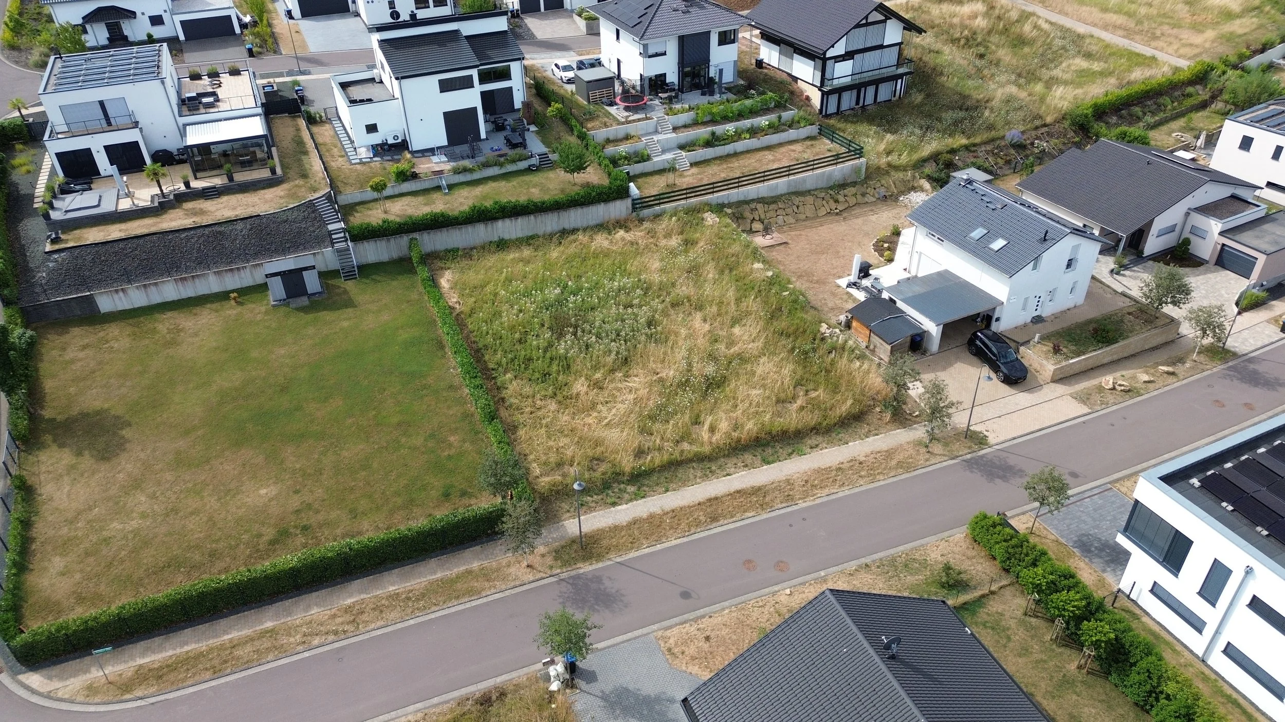 Vue aérienne d'un quartier résidentiel avec plusieurs maisons modernes, jardins et espaces verts, rue bordée d'arbres, et une zone de terrain vague au centre.