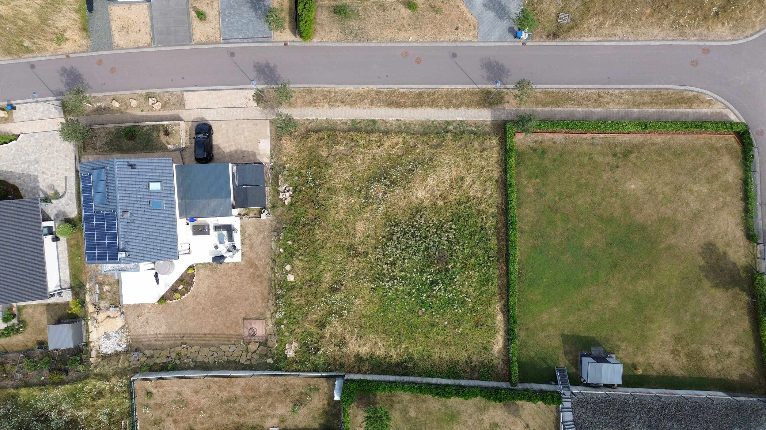 Balcon ou terrasse en haut, maison avec panneaux solaires sur le toit, jardin clôturé avec pelouse, allée pavée, petites haies, et espace végétal naturel.