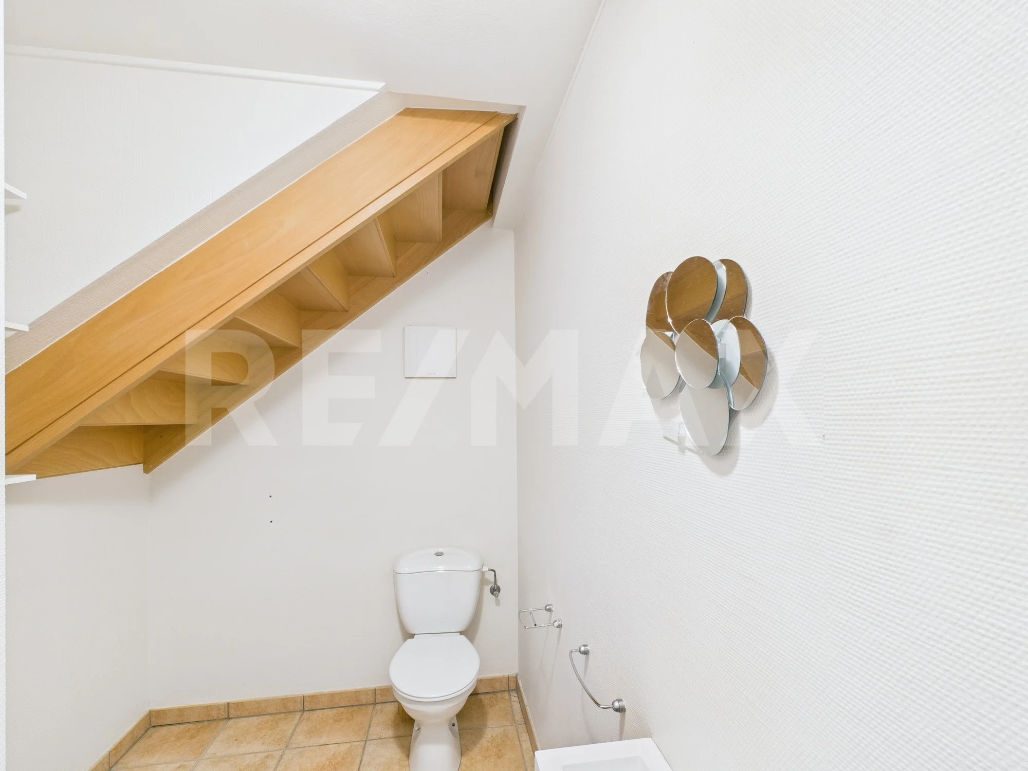 Petite salle de bain avec un WC blanc, un mur blanc, et une décoration murale métallique en forme de cercles. Un plafond en pente avec une étagère en bois.