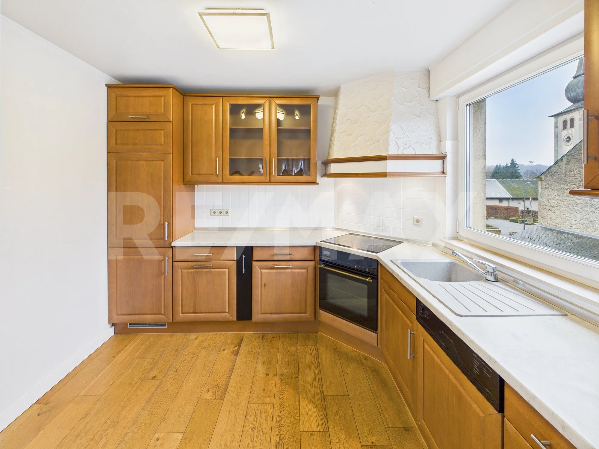 Cuisine avec des armoires en bois, une cuisinière, un évier près d'une grande fenêtre, plancher en bois, murs blancs.