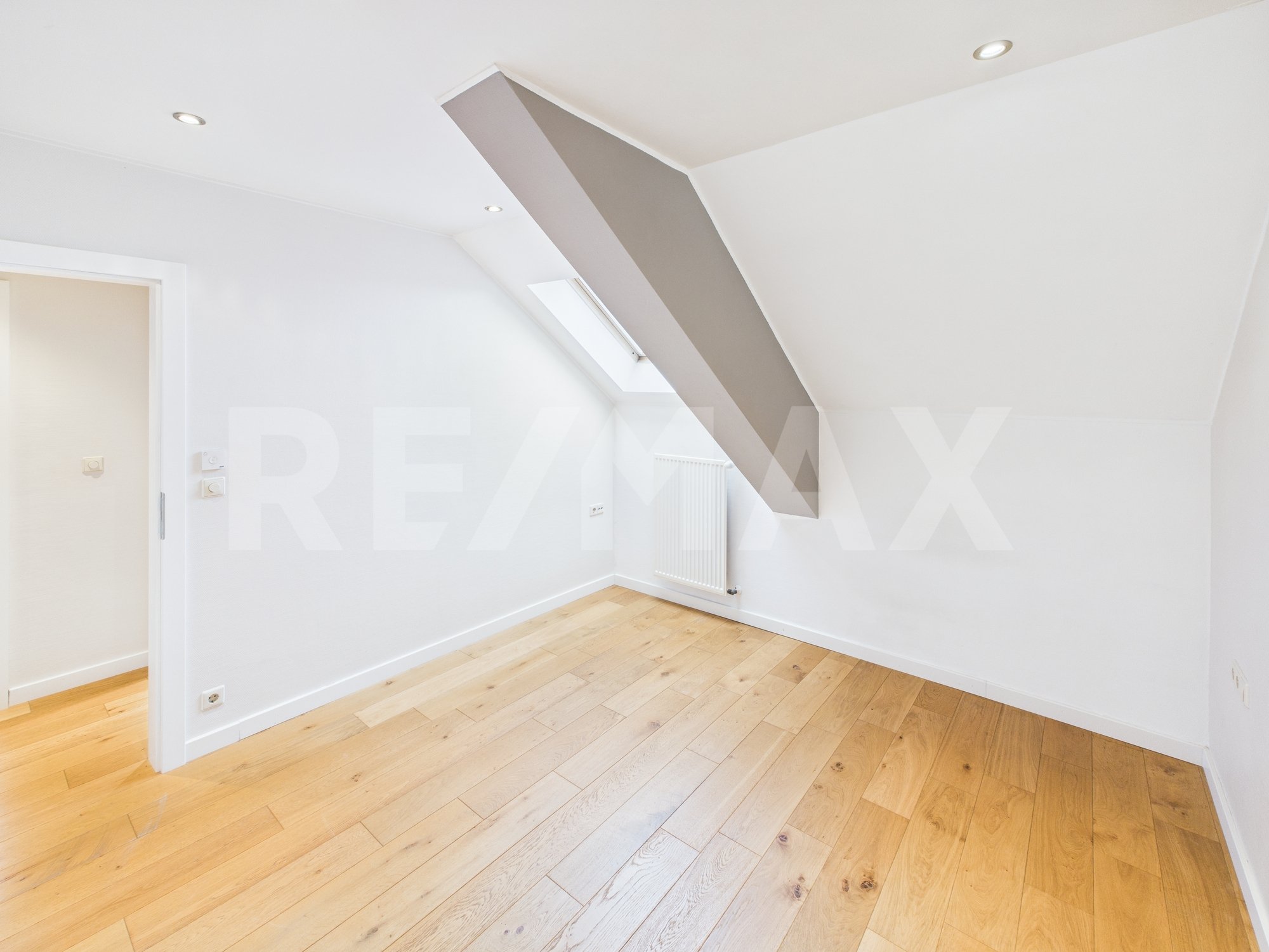Pièce vide avec murs blancs, plafond en pente, fenêtre de toit, radiateur, et plancher en bois clair.