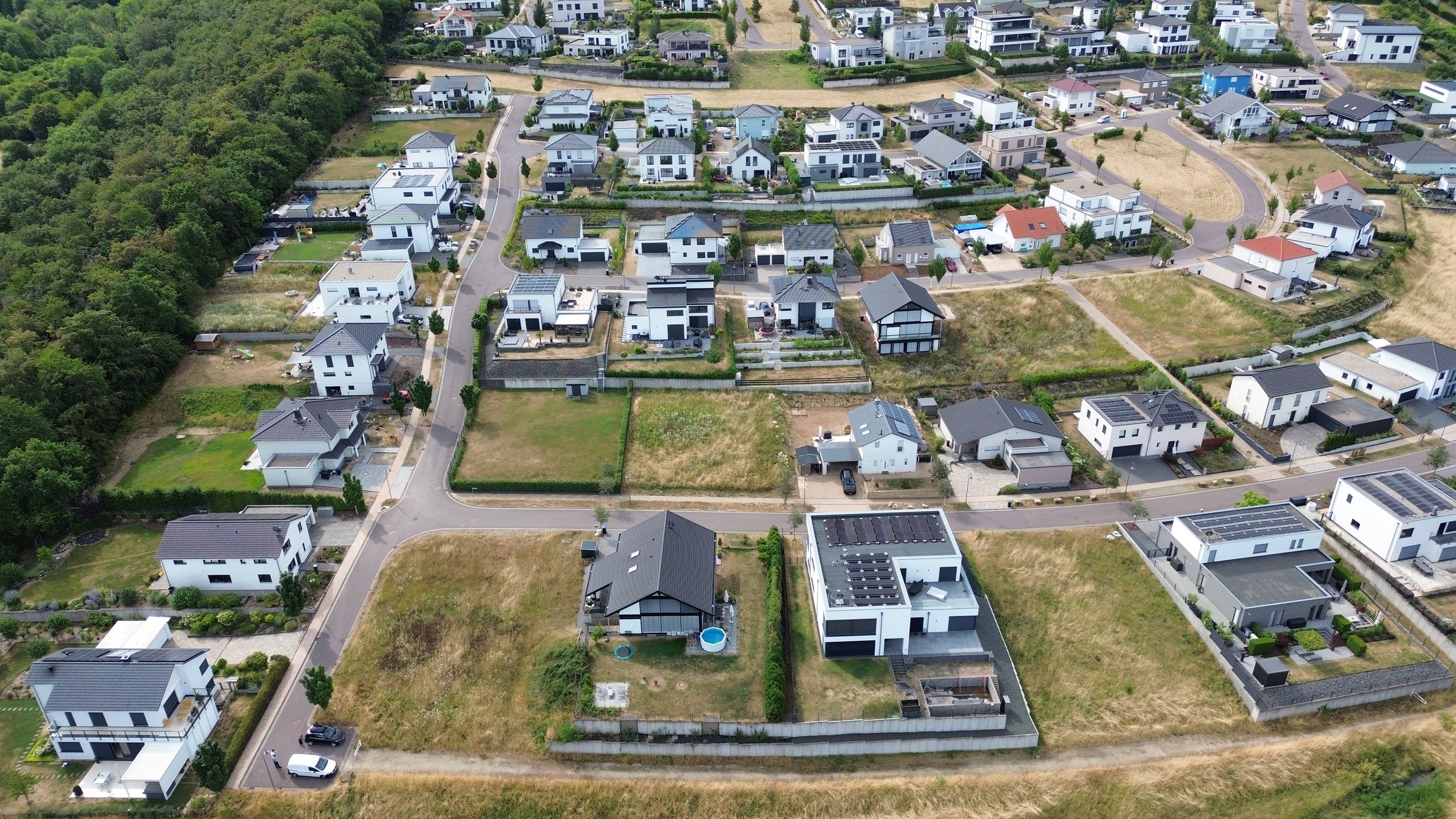 Vue aérienne d'un lotissement résidentiel avec des maisons modernes, des routes et des jardins, entouré de zones boisées et de terrains en développement.