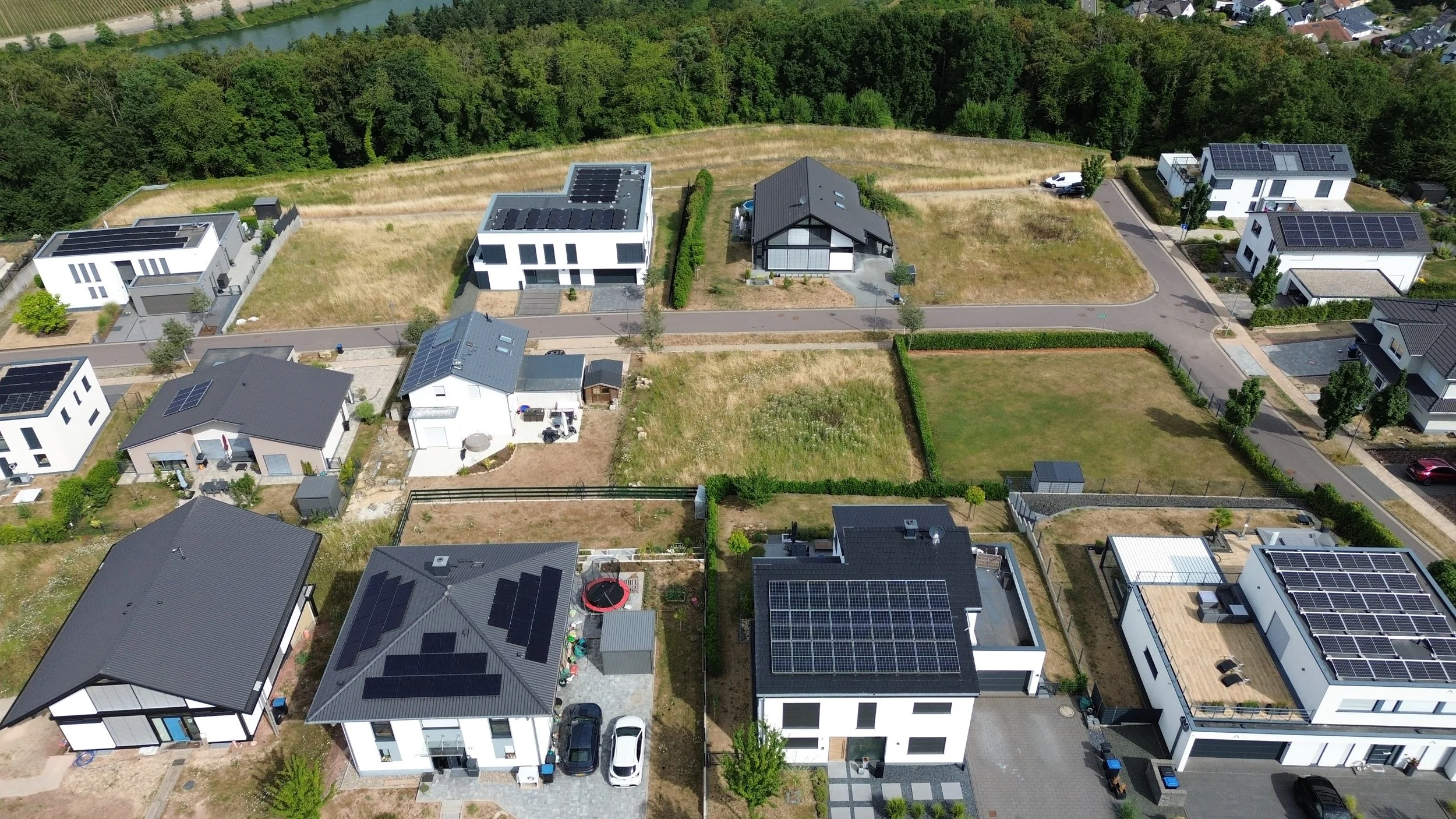 Vue aérienne d'un quartier résidentiel moderne avec des maisons équipées de panneaux solaires sur les toits, entouré de terrains verts, d'une forêt et d'une rivière.