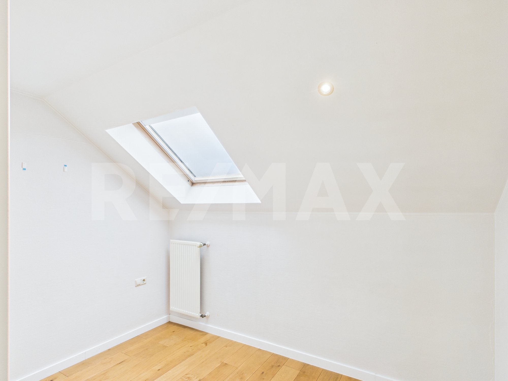Pièce avec un plafond incliné, un trou pour une fenêtre de toit laissant entrer la lumière naturelle, un radiateur blanc, un sol en bois clair, une prise électrique et un interrupteur.