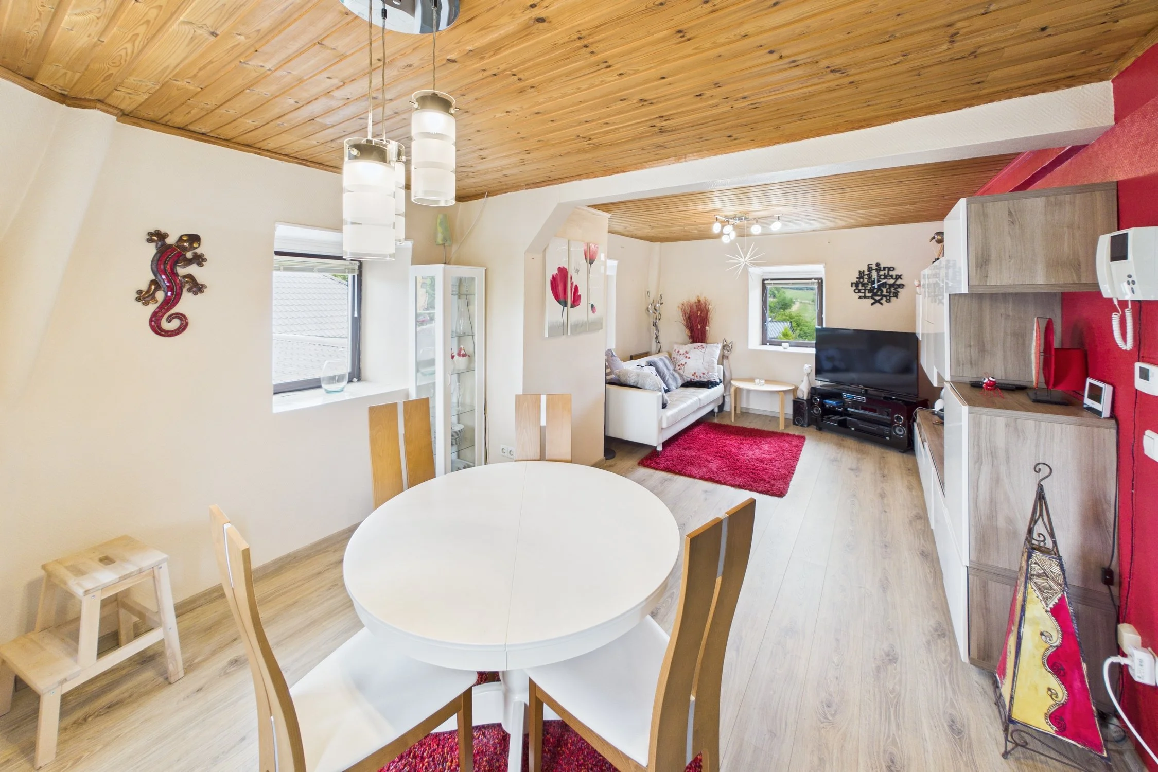 Salon de salon avec une table ronde blanche, chaises en bois, canapé beige, téléviseur, étagère en bois, tapis rouge, murs blanc et rouge, fenêtres avec vue sur un jardin, plafond en bois.