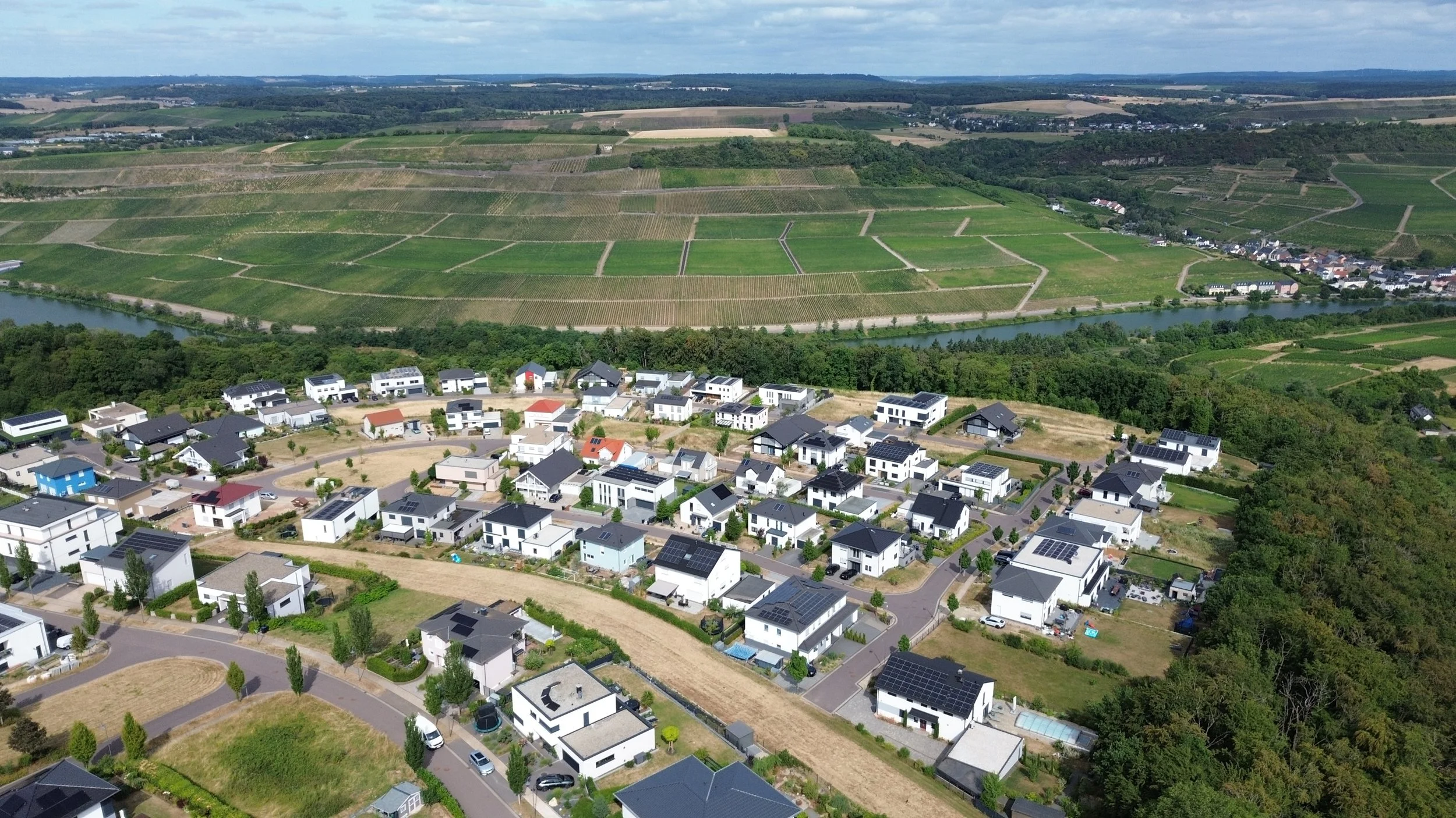 Vue aérienne d'une banlieue résidentielle avec maisons modernes, routes, un fleuve et des paysages agricoles verdoyants au fond.