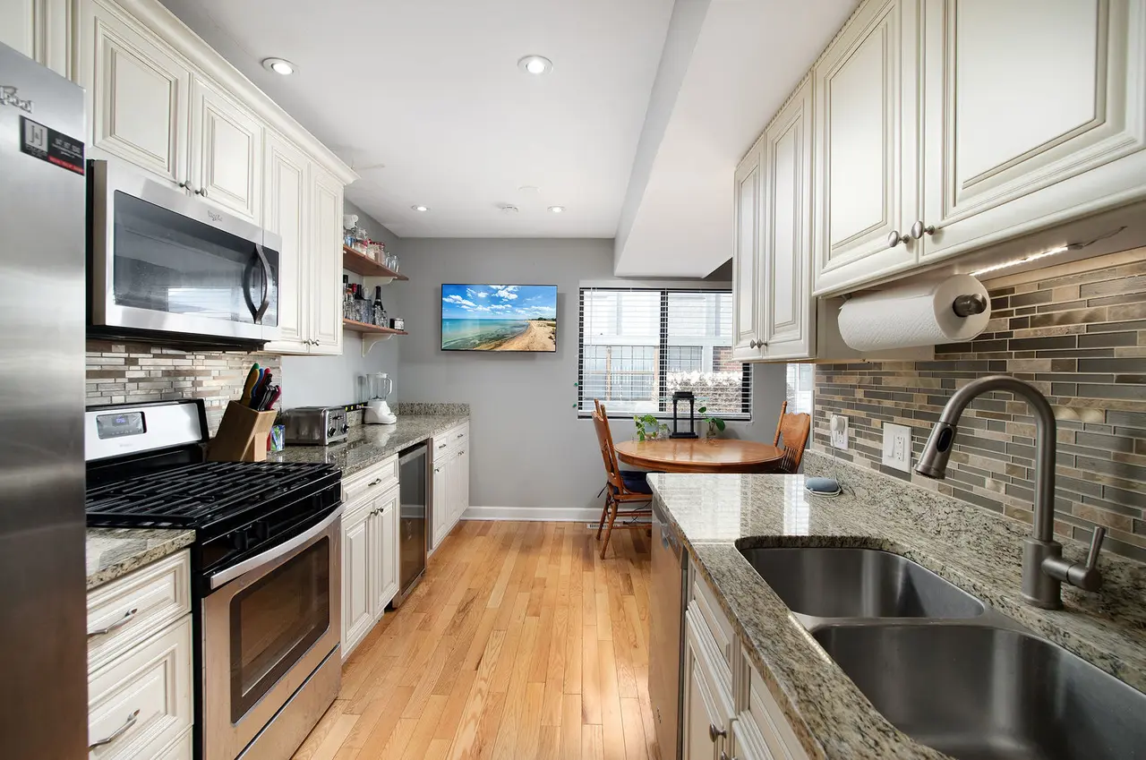 Kitchen with white cabinets, granite countertops, stainless steel appliances, a small dining table with three chairs, a window with blinds, and a TV on the wall.