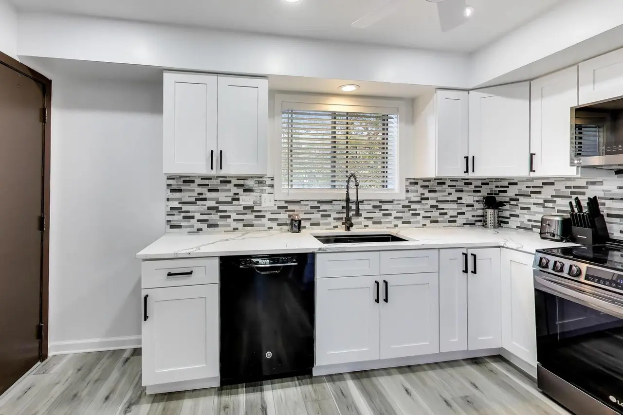 Modern kitchen with white cabinets, a black dishwasher, and a black stove. Gray, black, and white mosaic tile backsplash, window above the sink, white marble countertops, and wood-style flooring.