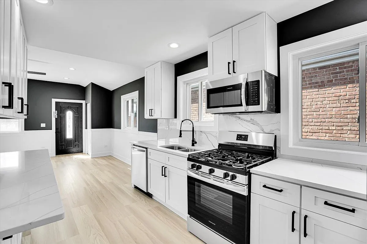 Modern kitchen with white cabinets, black handles, stainless steel appliances including microwave and gas stove, white countertops, a black faucet, and windows overlooking a brick wall outside.