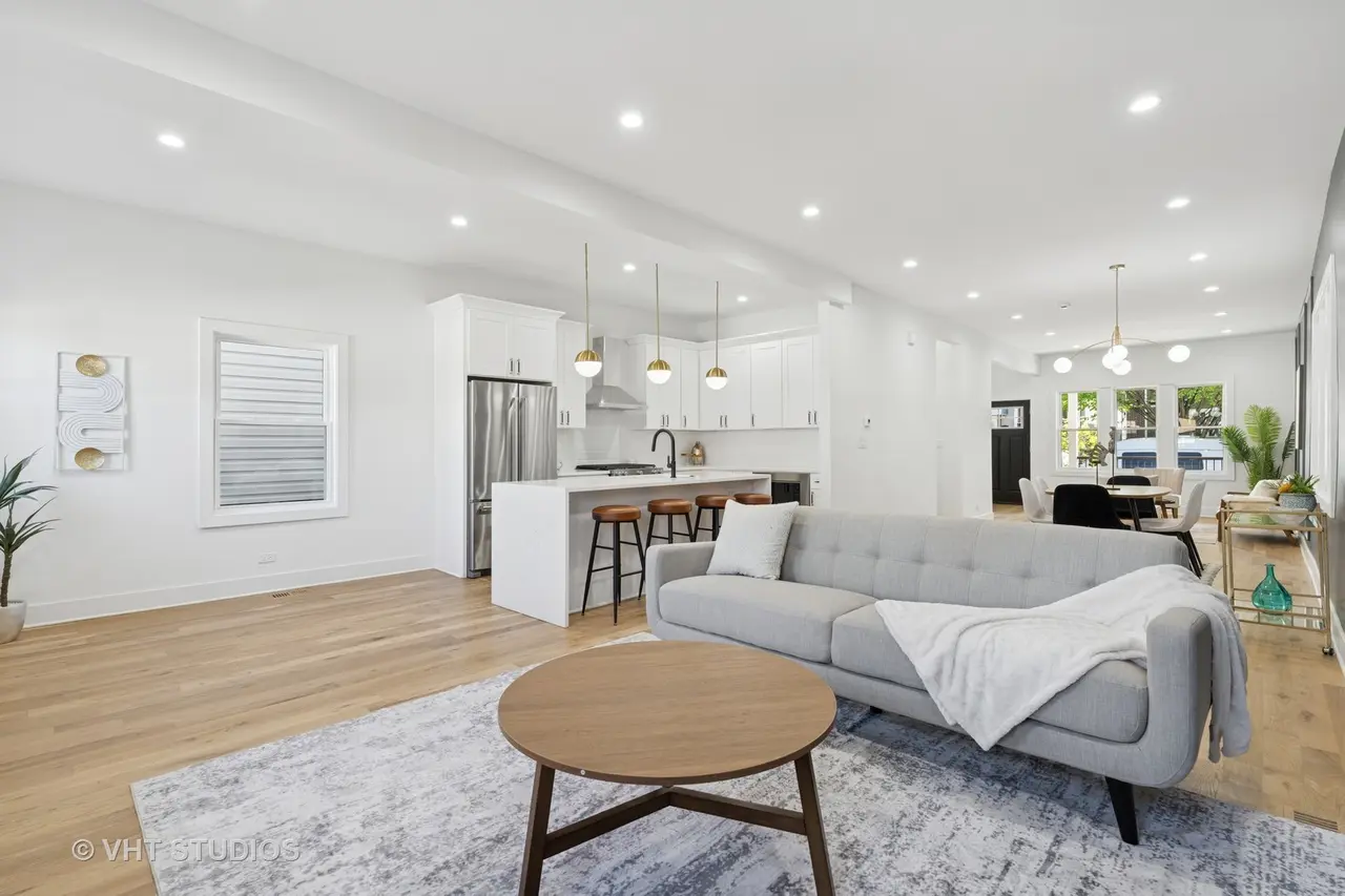 Bright, modern open-concept living room and kitchen with white walls, hardwood floors, gray sofa, round wooden coffee table, kitchen island with barstools, and dining area with round table and white chairs.