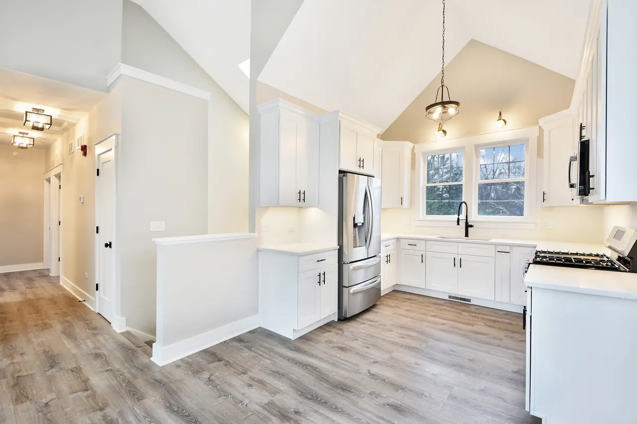 Bright kitchen with white cabinets, stainless steel refrigerator, and a window above the sink, featuring a wood floor and ceiling lights.