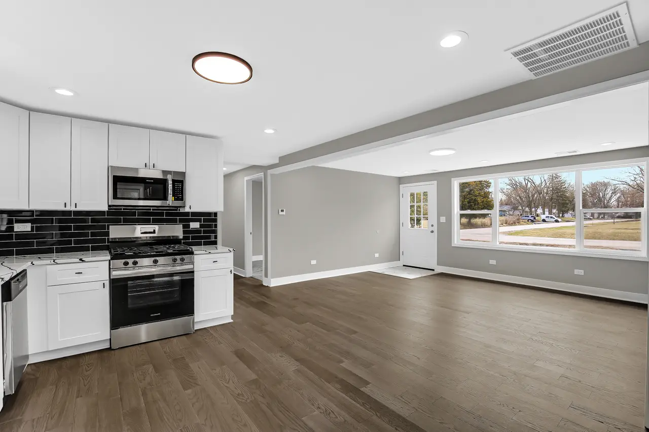 Empty living room and kitchen area in a modern home with large windows, light gray walls, hardwood flooring, white cabinets, and stainless steel appliances.