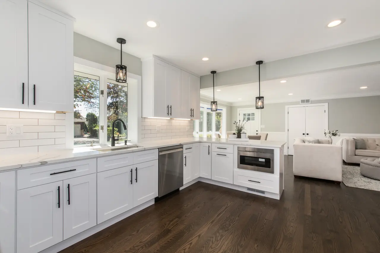 Modern kitchen with white cabinets, dark hardwood floors, stainless steel appliances, and black pendant lights over the island, open to a living area with white furniture.