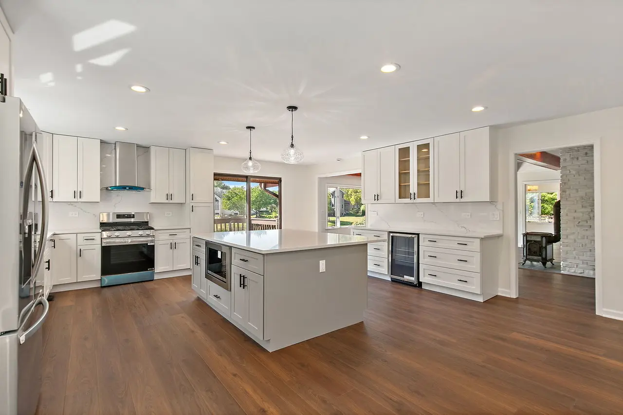 Modern kitchen with white cabinets, stainless steel appliances, a central island, and hardwood floors, with large windows showing an outdoor backyard.