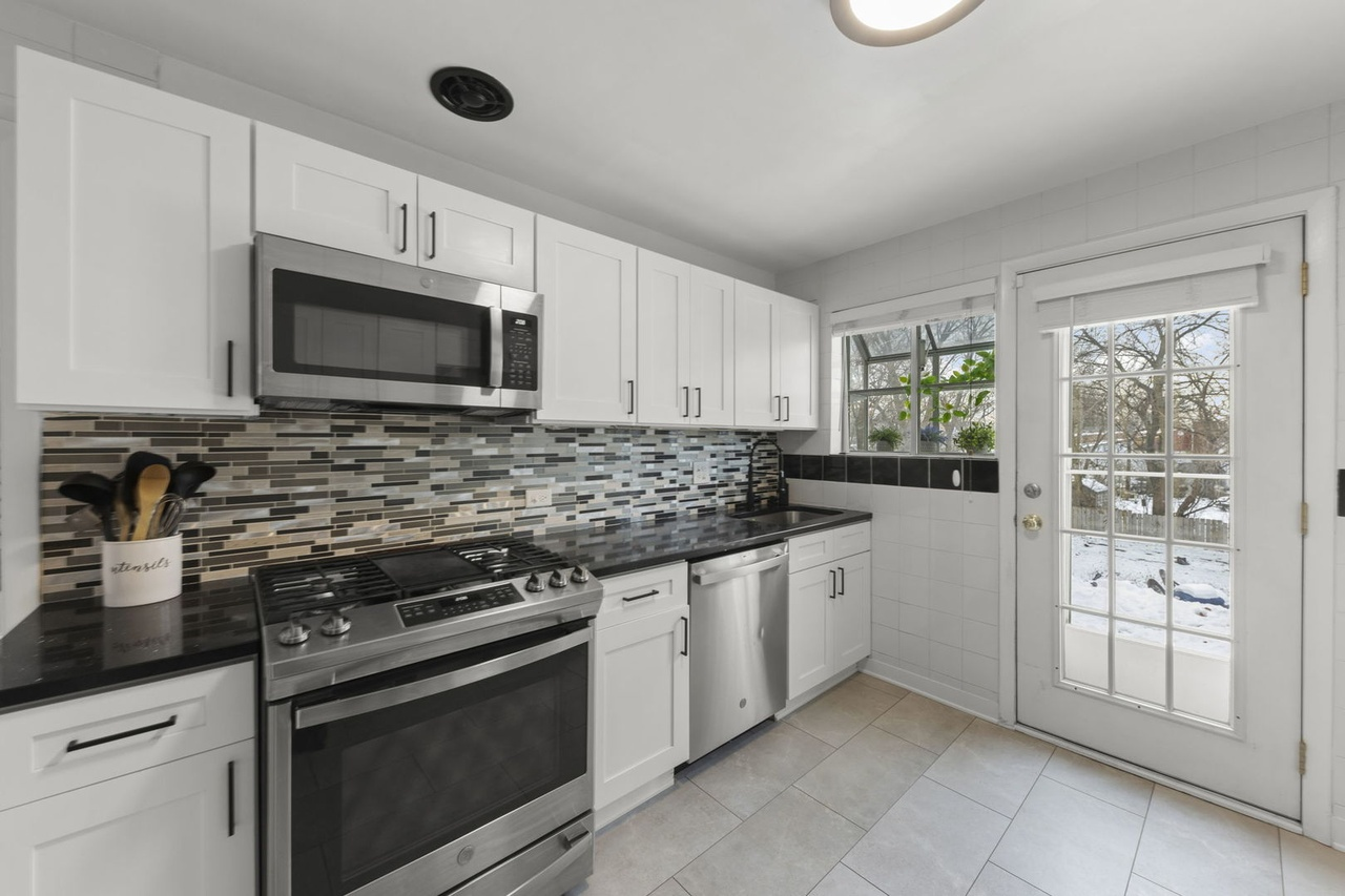 White kitchen with stainless steel microwave, stove, and dishwasher. Black countertop, mosaic tile backsplash, and a window with plants. Outside shows snow and trees.