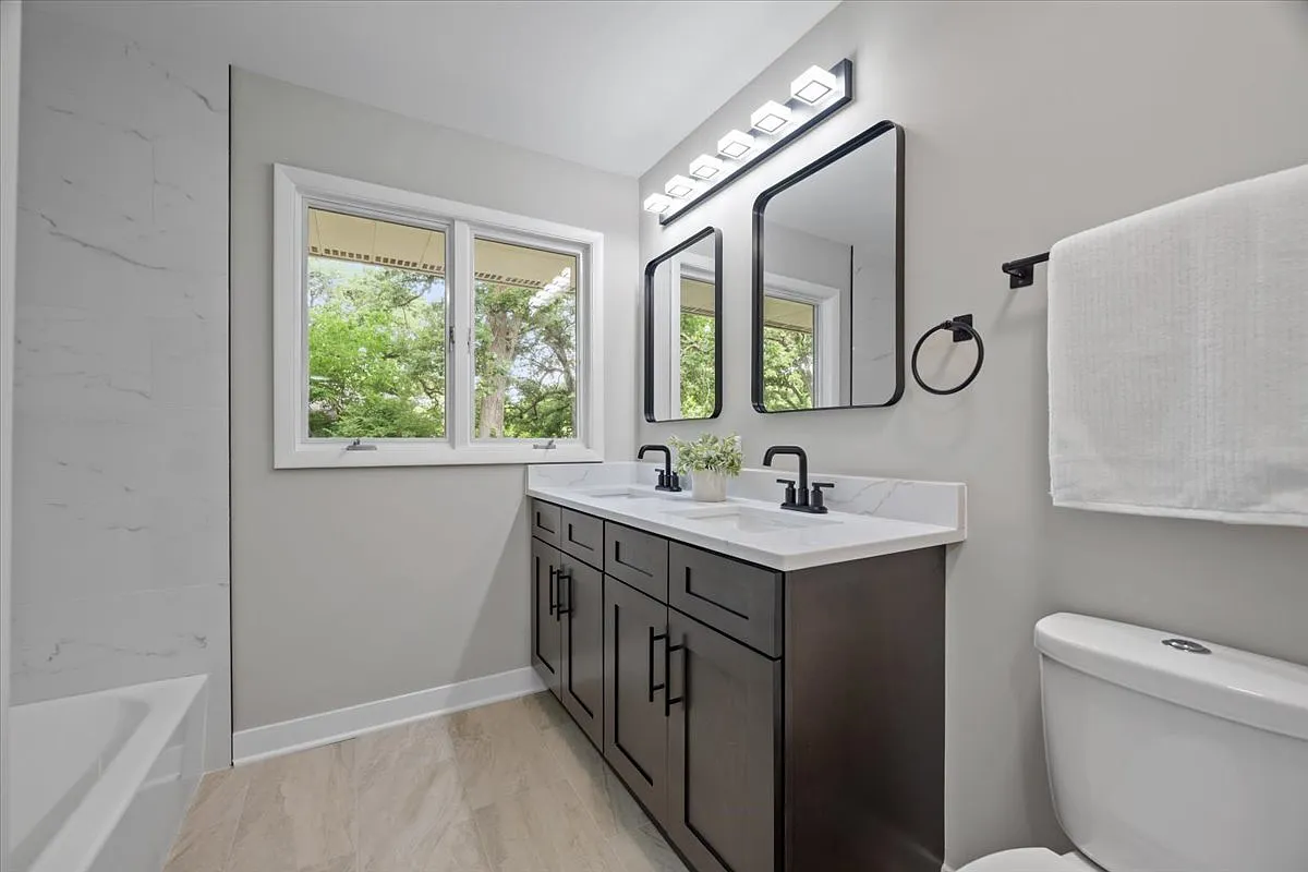 Modern bathroom with double vanity, black faucets, two mirrors, towel ring with towel, window showing trees outside, and beige tile flooring.