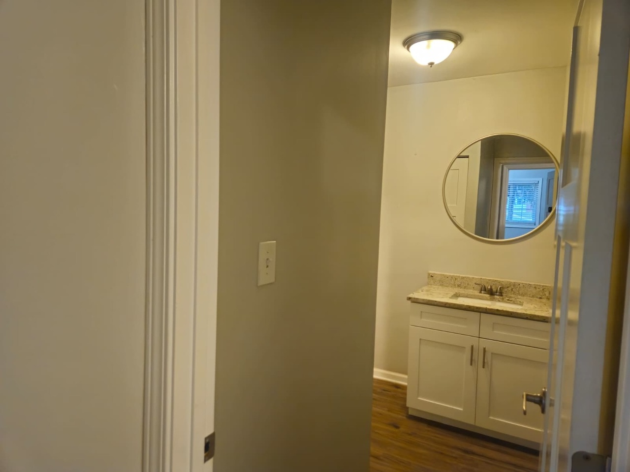 Interior view of a bathroom with a white vanity, granite countertop, round mirror, and window, seen from a hallway with beige walls and hardwood floors.
